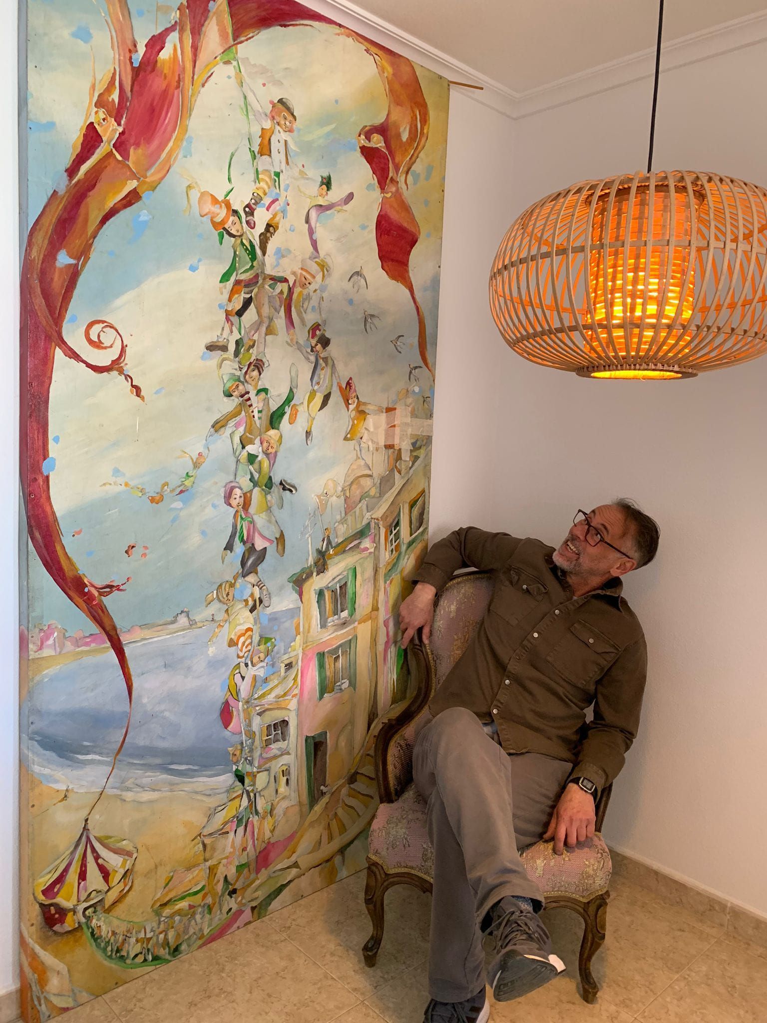 Persona recostada en una silla junto a un colorido mural y una lámpara colgante de mimbre en el interior.