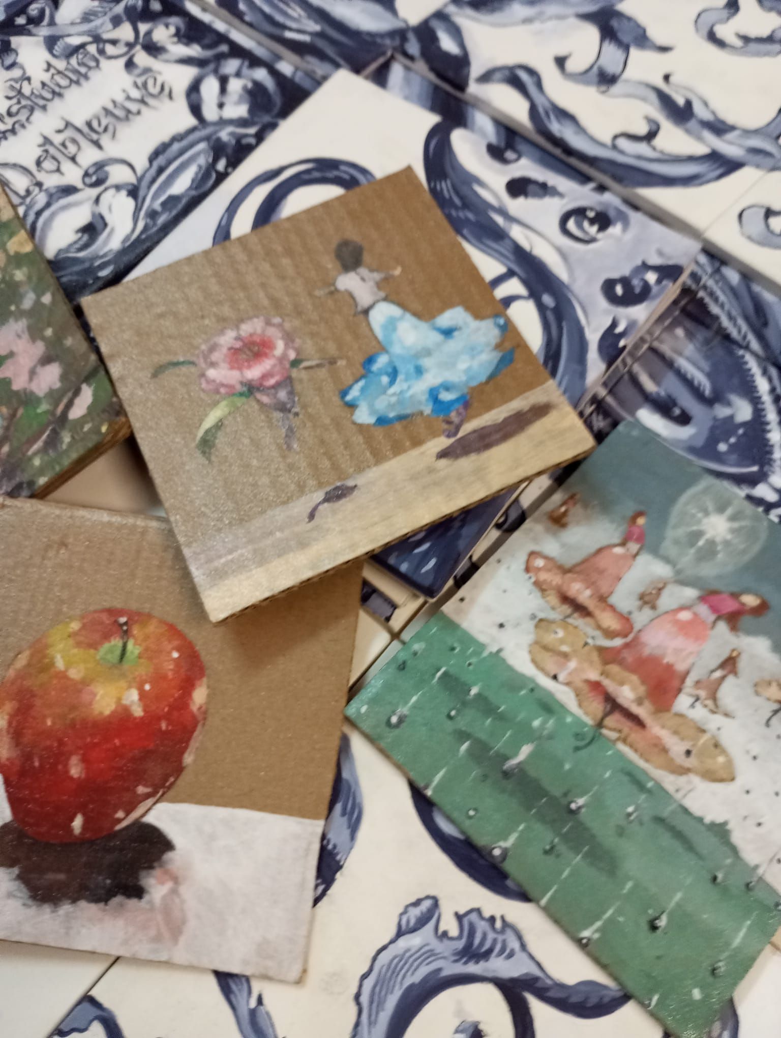 Tres pequeños cuadros sobre papel: una fruta roja, flores azules y una escena en tonos pastel sobre tela estampada.