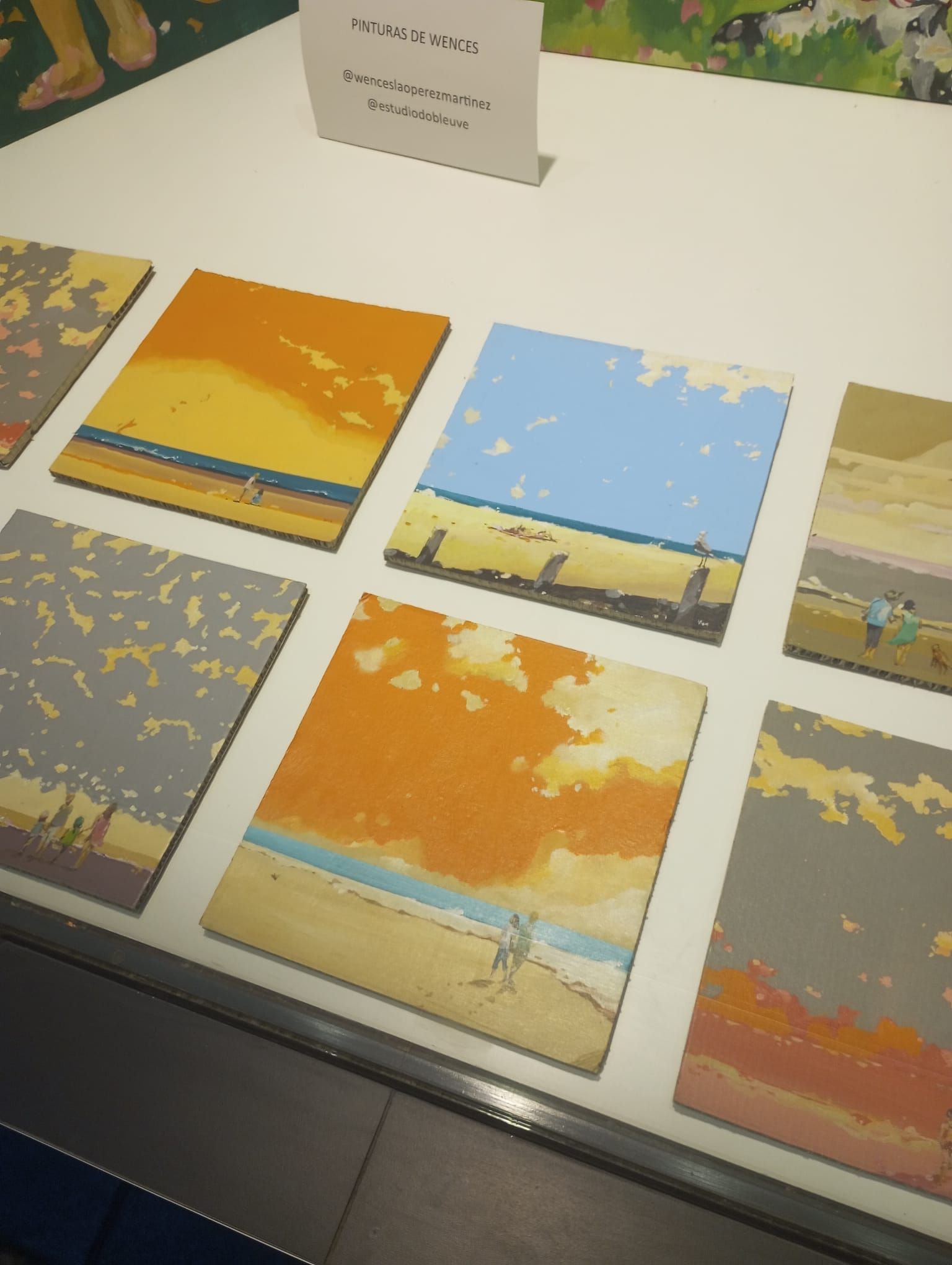 Una mesa de exposición con coloridas láminas de paisajes en tonos amarillos, azules y grises, además de un pequeño cartel.