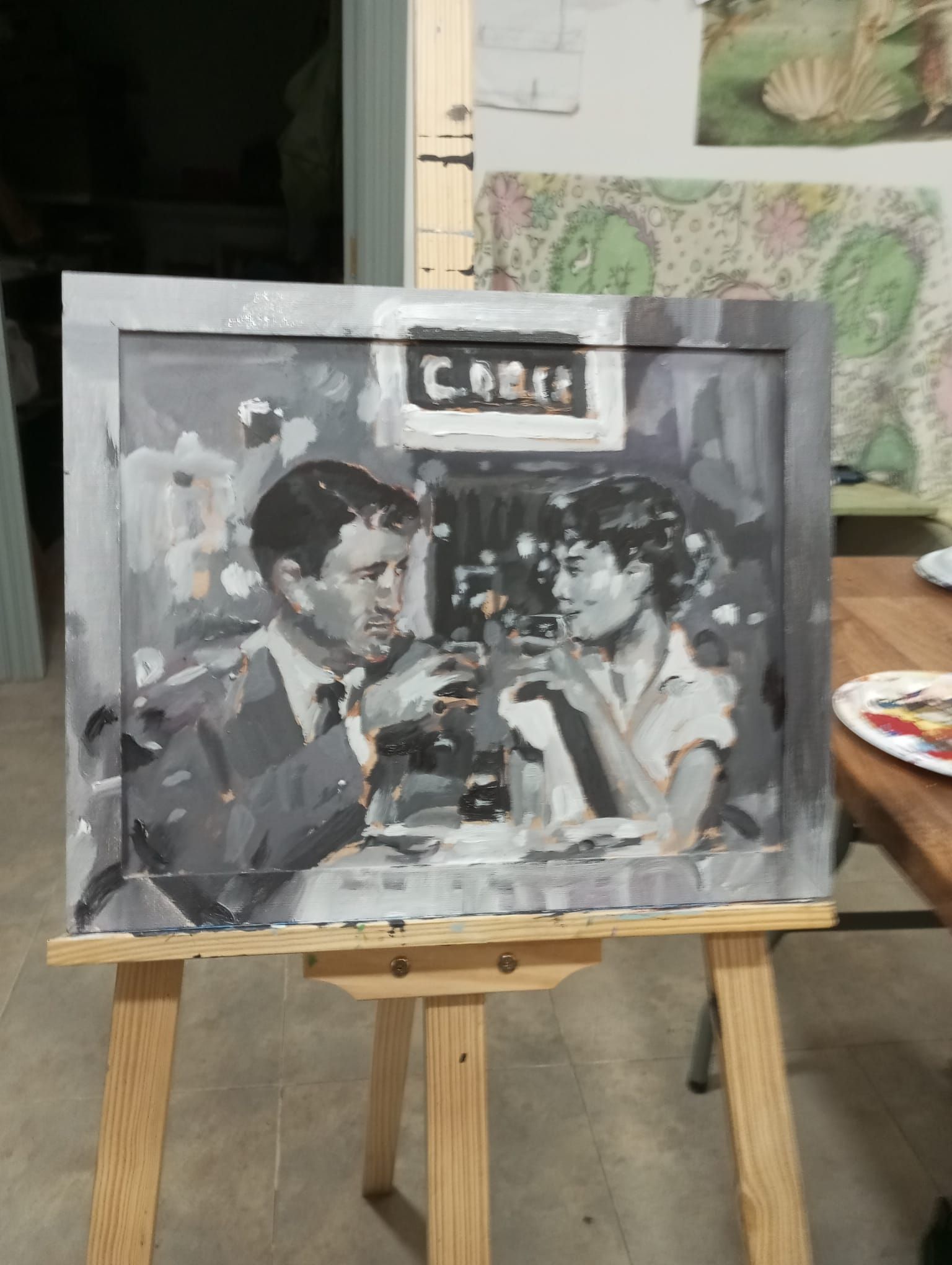 Cuadro en blanco y negro de dos personas conversando alrededor de una mesa, sobre un caballete, en un estudio de arte.