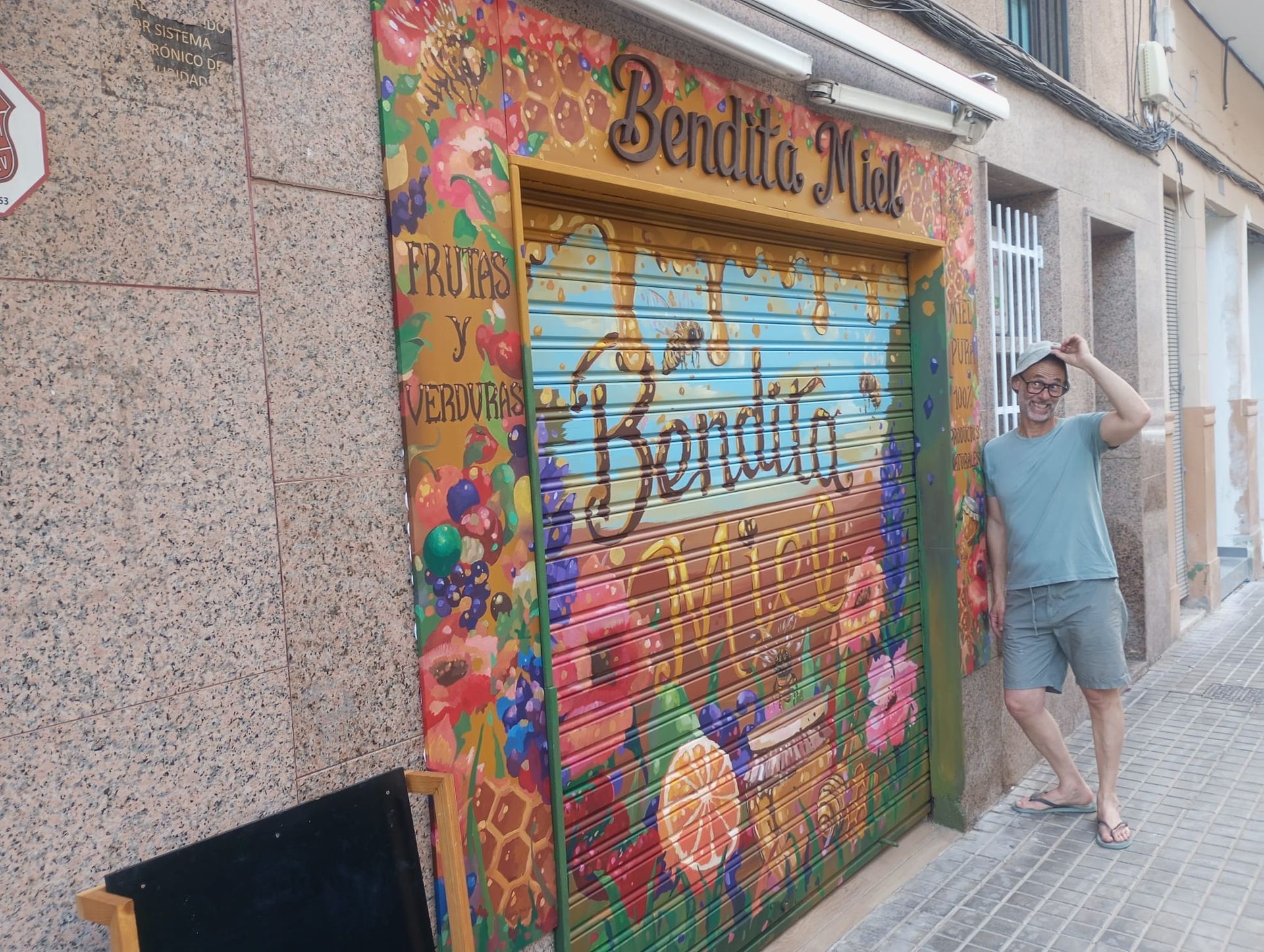 Persona posando junto a la fachada de una tienda, cubierta de coloridos grafitis, con una persiana azul y una puerta enrollable.