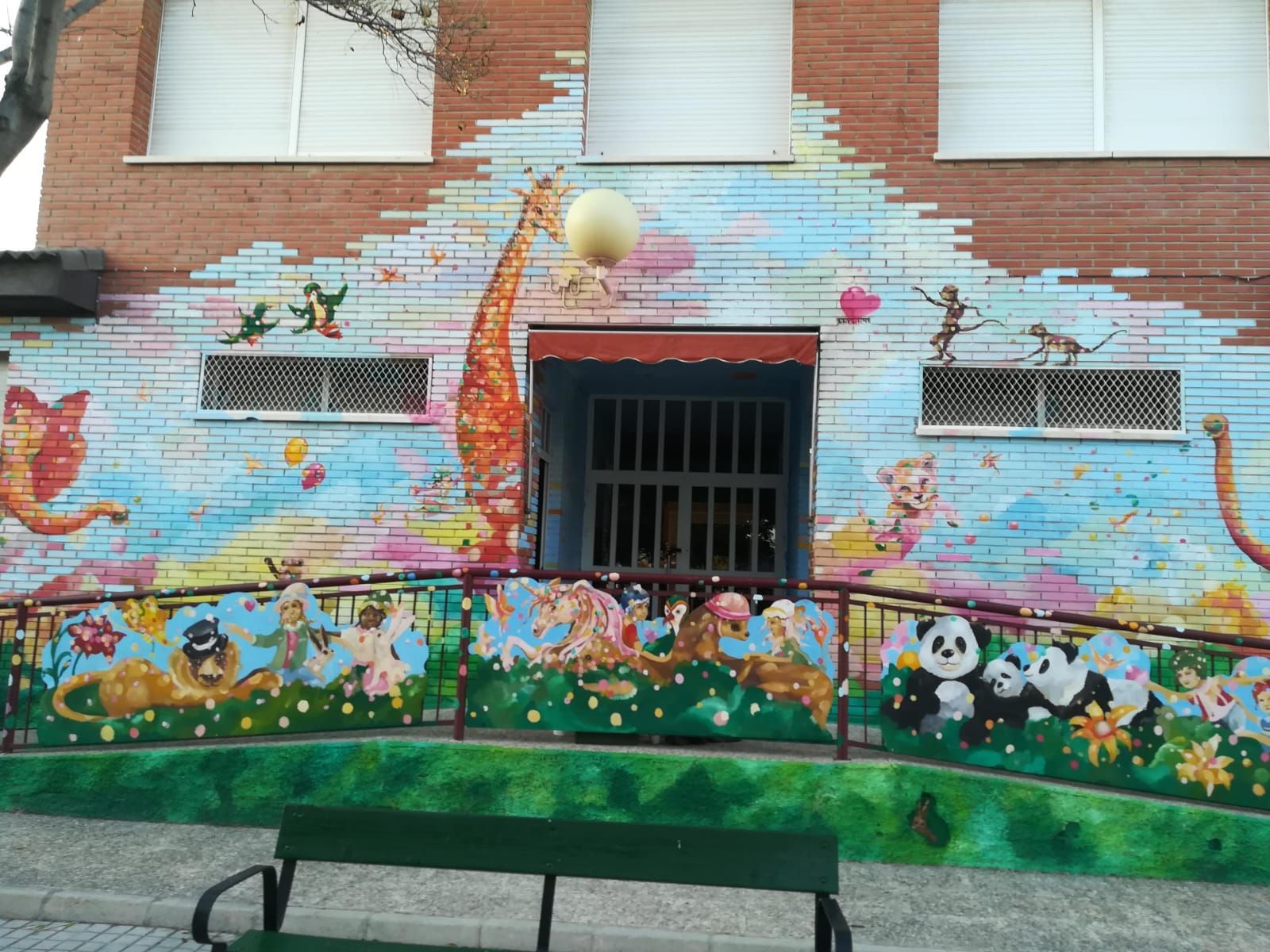 Mural colorido en una pared de ladrillo con flores y pandas alrededor de una ventana enrejada y un banco verde.