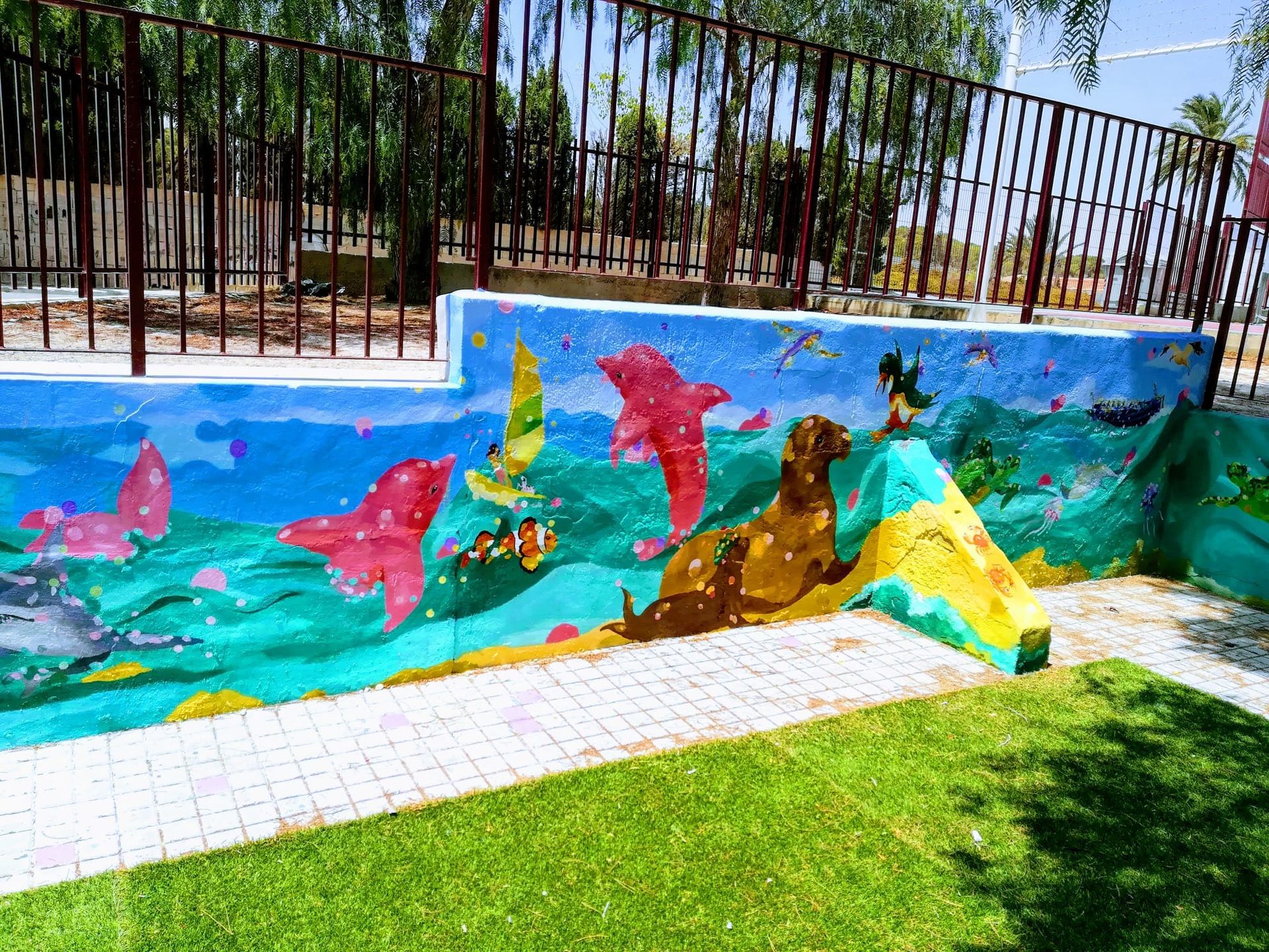 Un colorido mural de peces y vida marina pintado en una pared exterior junto a un sendero cubierto de hierba.