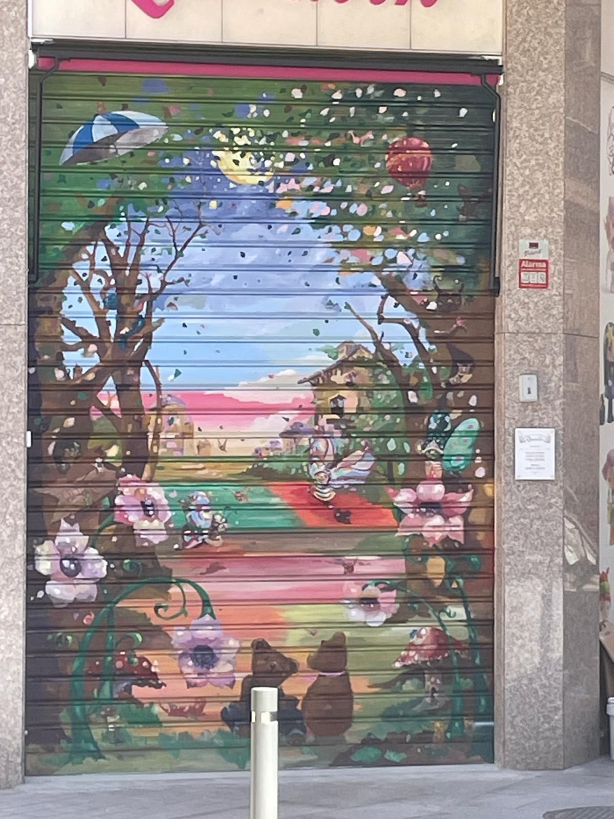 Mural colorido en la persiana de una tienda con un sendero, árboles, flores y una escena de una puesta de sol rosada.