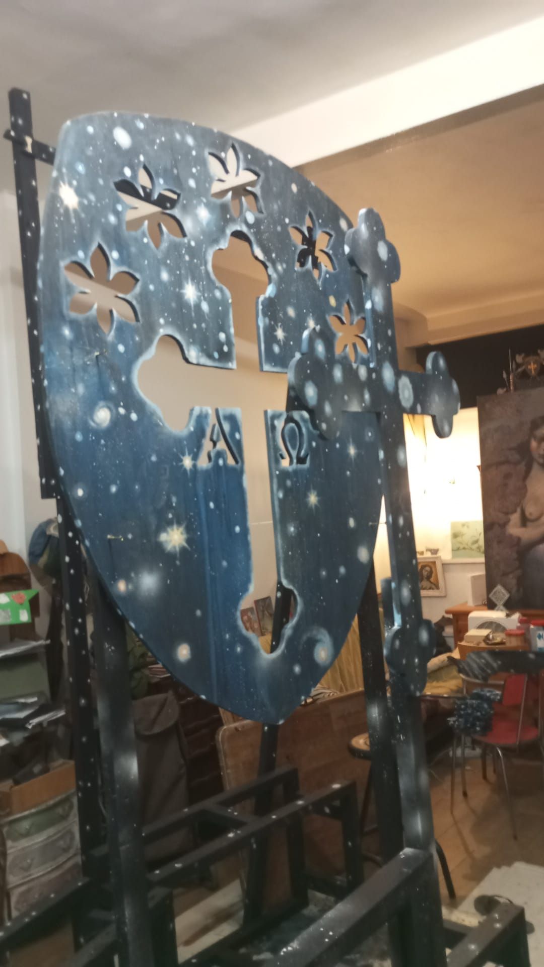 Escudo azul con una cruz blanca y recortes de estrellas, expuesto en el interior sobre un soporte.