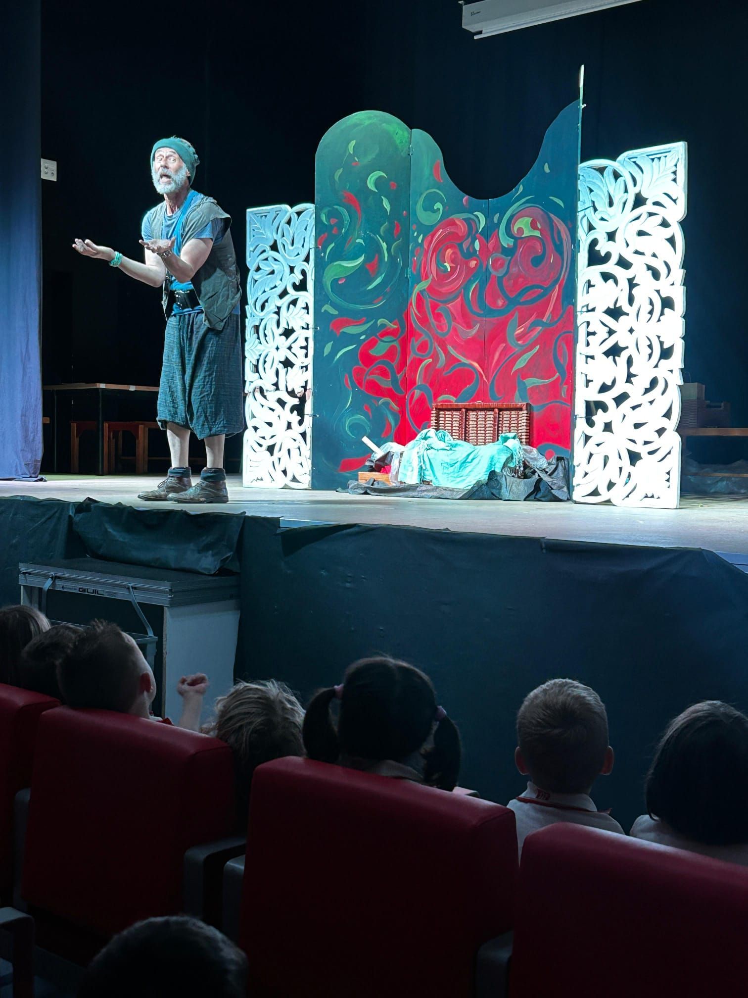 El orador se encuentra en el escenario, junto a coloridos paneles abstractos, dirigiéndose a un público sentado en butacas rojas de teatro.