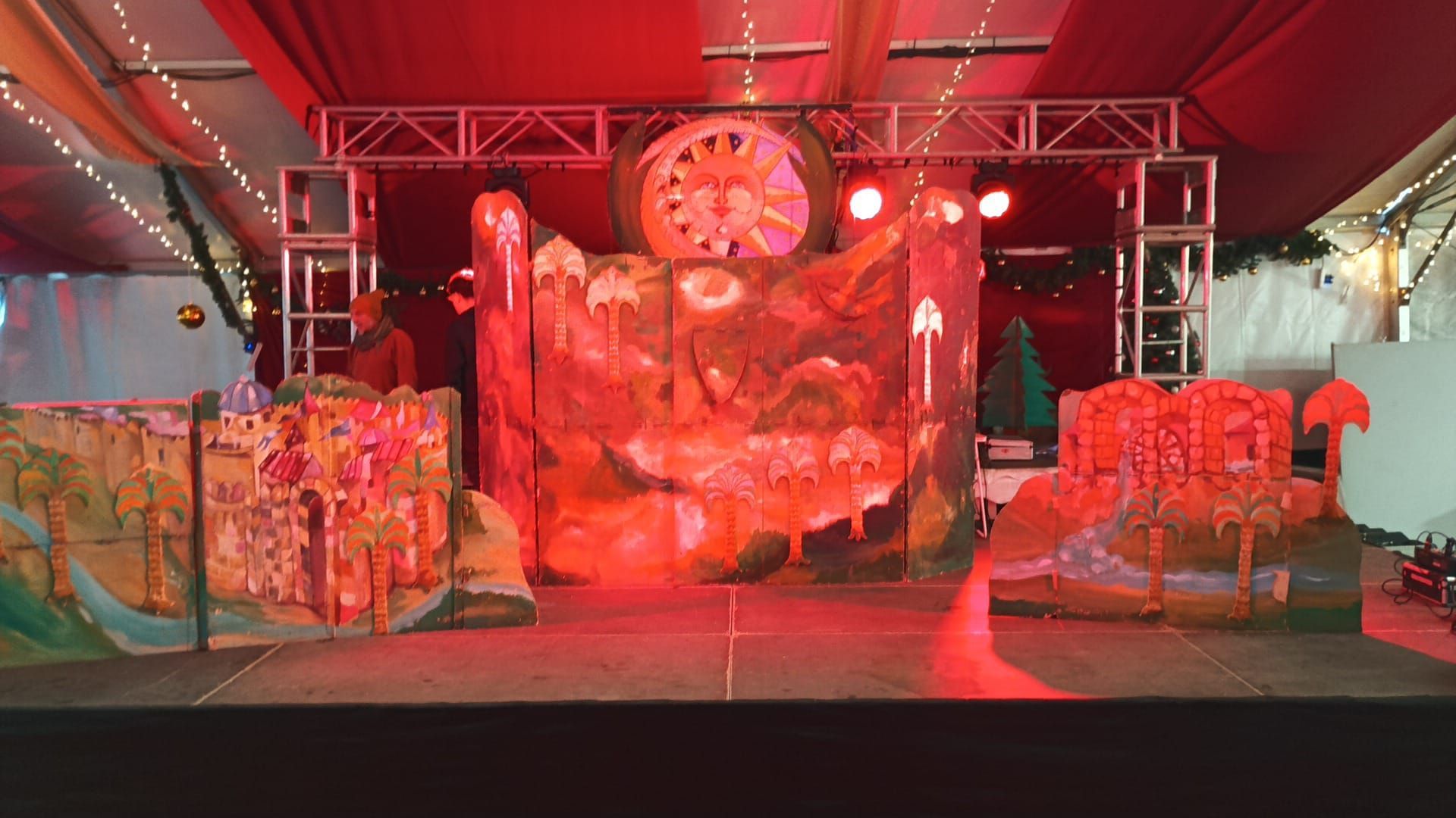 Escenario iluminado en rojo con un gran reloj como telón de fondo, paneles coloridos y accesorios bajo una cubierta que simula una carpa.