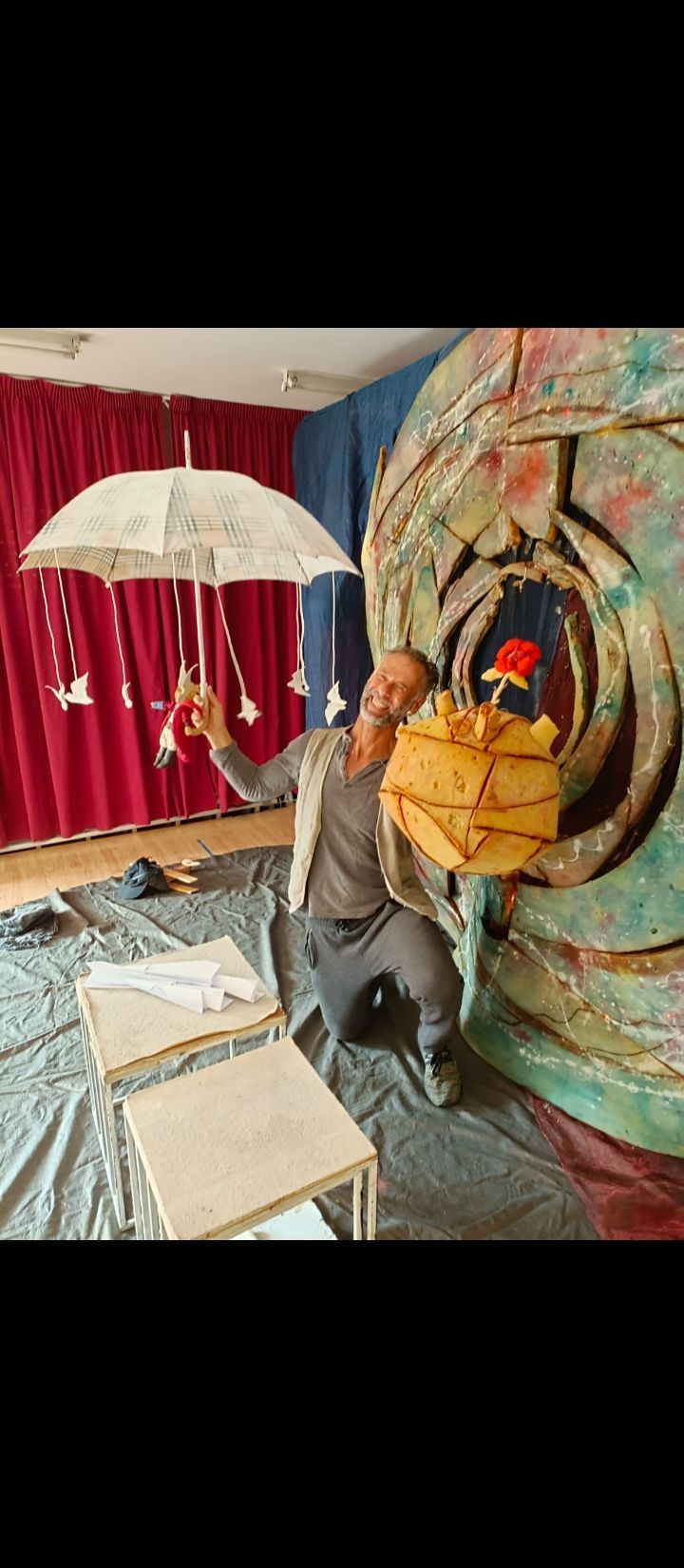Un hombre con una sudadera gris posa junto a un colorido mural de estilo circense y una cortina roja en el interior de un edificio.