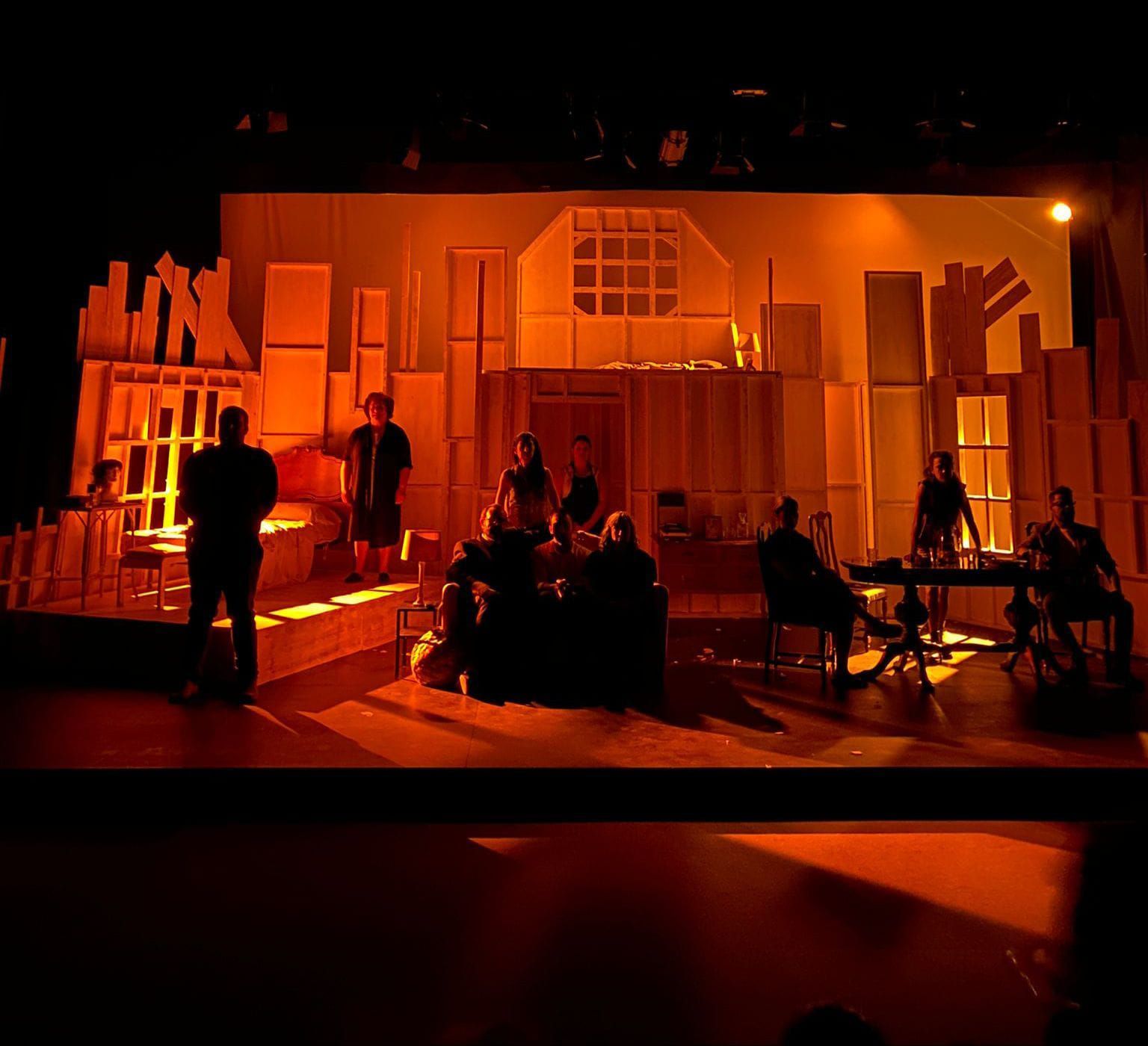 Escenario iluminado con luz naranja con varios artistas silueteados en un decorado teatral que se asemeja a habitaciones y edificios.