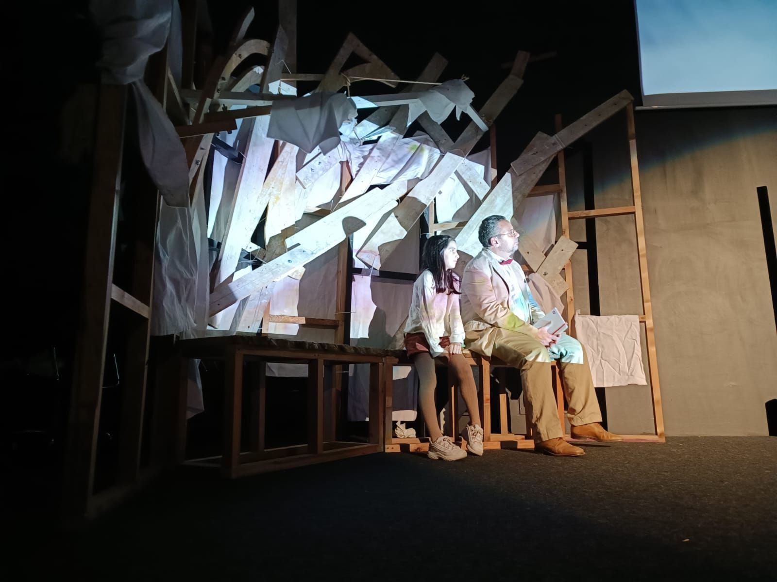 Dos personas sentadas en un escenario con un telón de fondo blanco angular y accesorios de madera bajo una iluminación cálida.