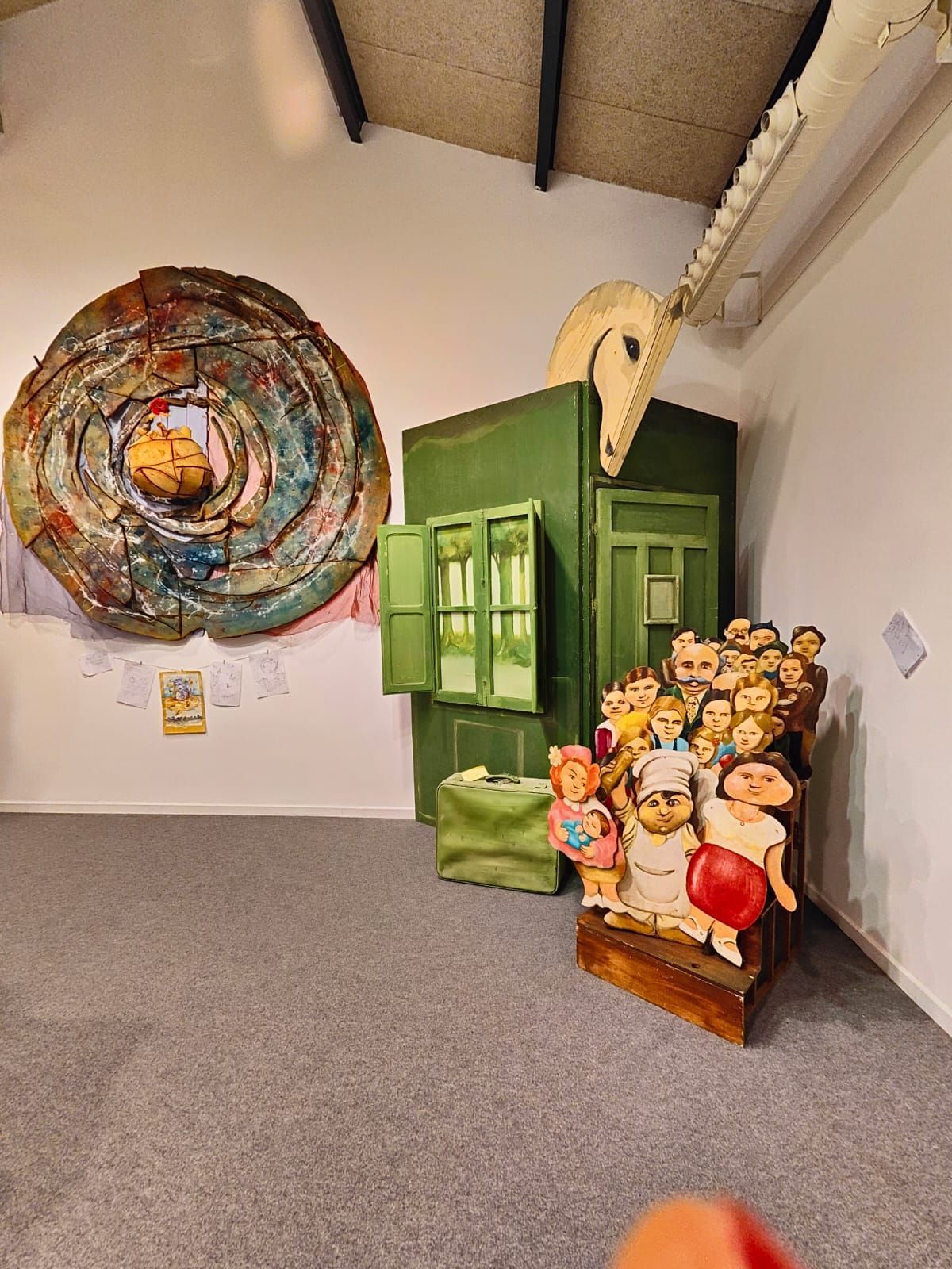 Rincón de galería con una colorida pieza circular en la pared, una escultura verde y un grupo de figuras de muñecas en el rellano de una escalera.