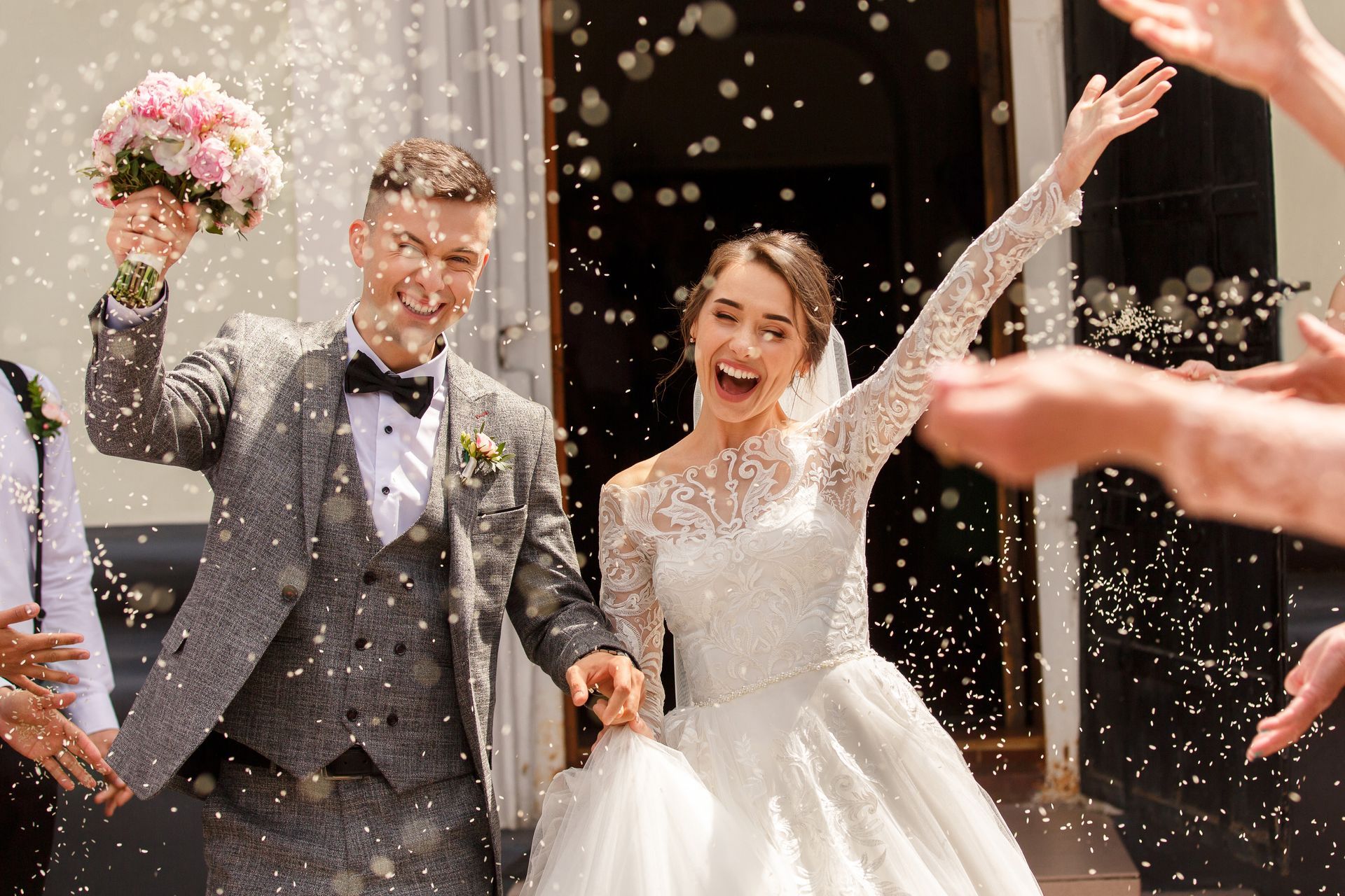 Les jeunes mariés célèbrent leur union en levant les bras et en lançant des confettis. La mariée, en robe, tient son bouquet, tandis que le marié porte un costume.