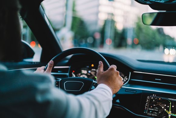 Les mains du conducteur sur le volant à l'intérieur d'une voiture moderne, avec une rue de la ville visible devant lui.