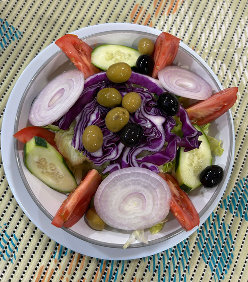Ensalada en un recipiente blanco: tomates, pepino, cebolla, aceitunas y repollo morado sobre una estera tejida.