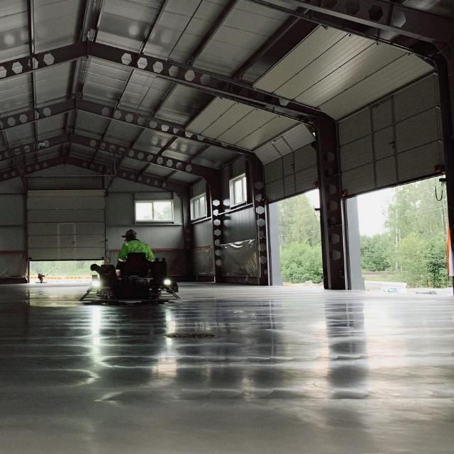 Työntekijä tasoittaa ajettavalla sähköhiertimellä kiillotettua betonilattiaa suuressa, avorakenteisessa varastossa.
