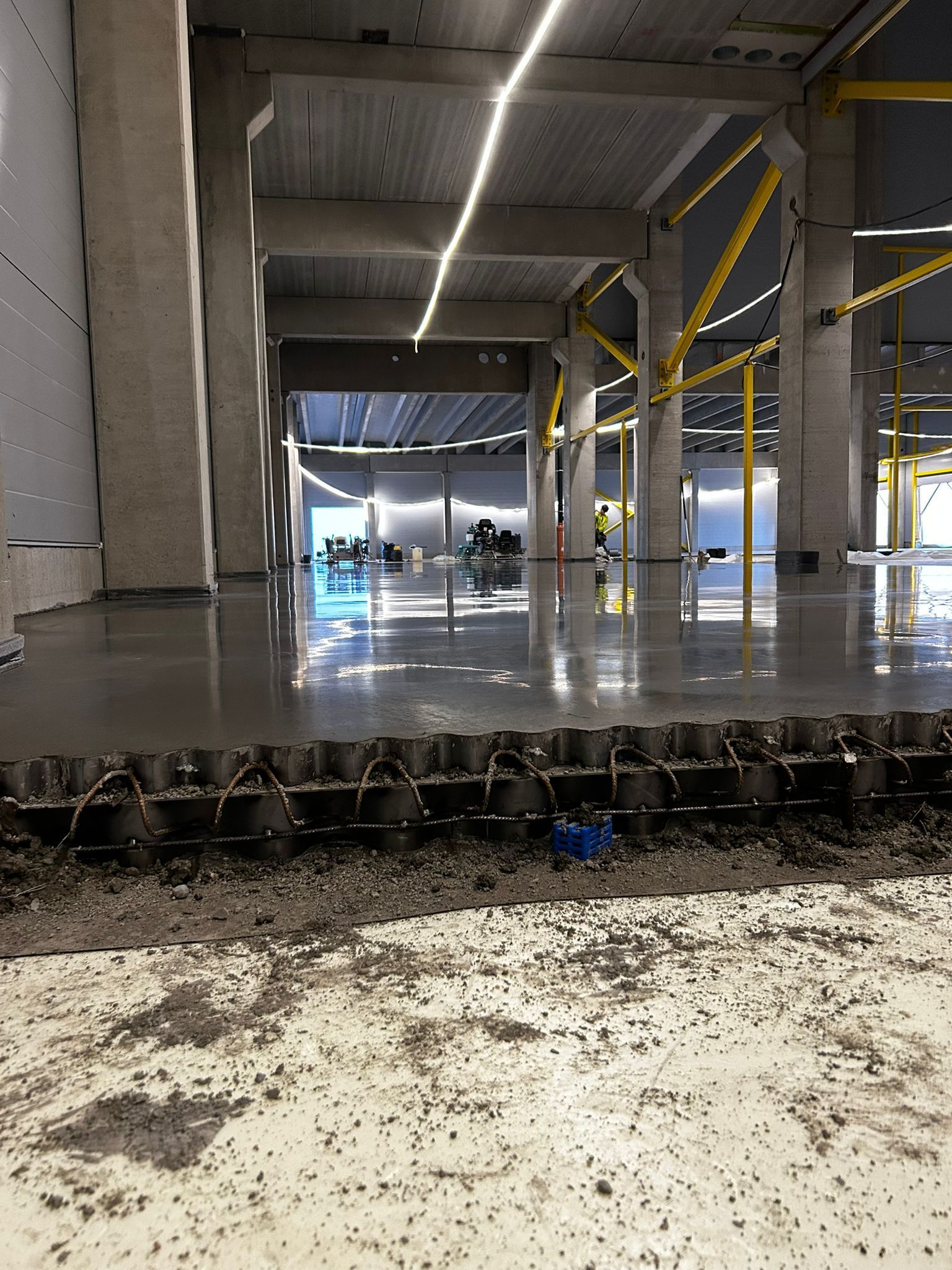 Rakennustyömaan sisätilat: betonilattia, pylväät ja kattovalaistus. Etualalla mustaa aaltopeltiä.