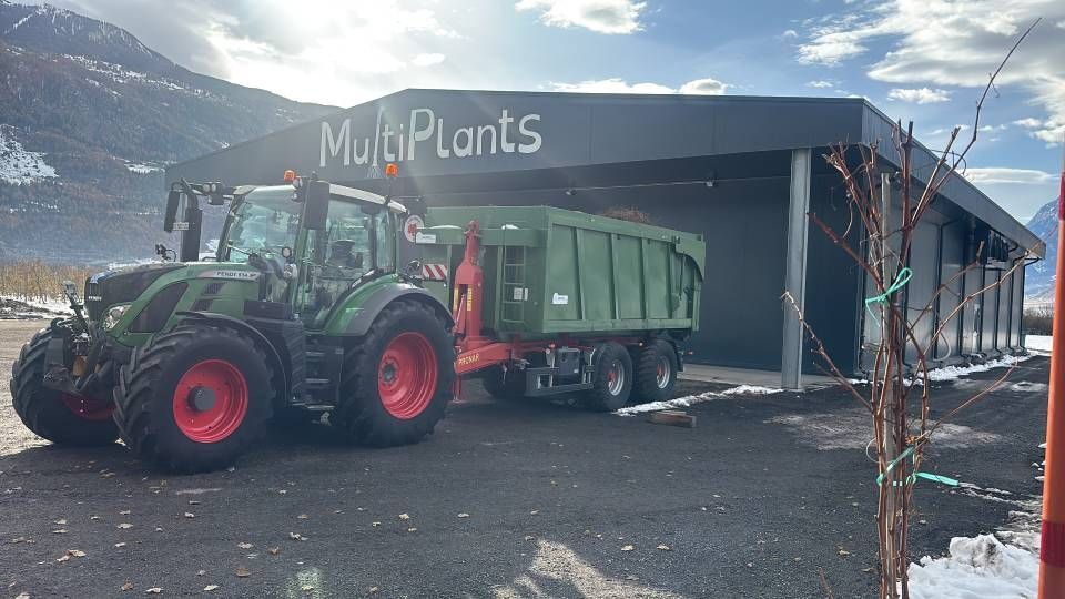 Un tracteur vert avec une remorque charge des matériaux dans l'usine MulciPlants, près des montagnes.