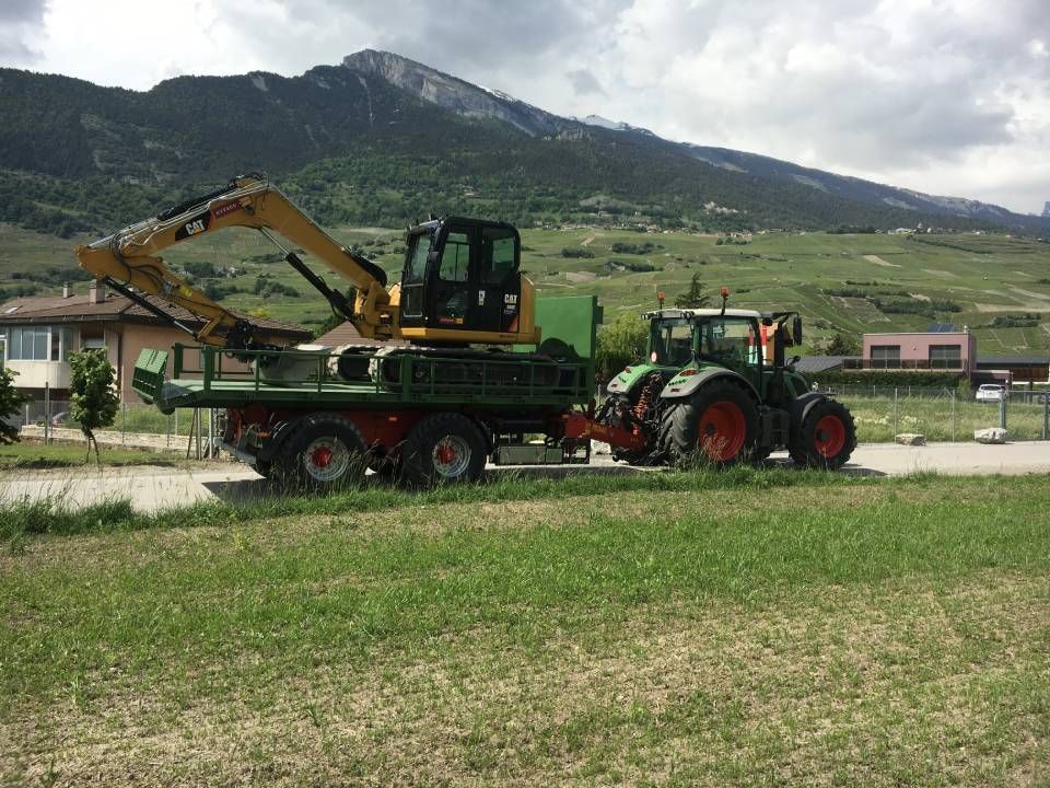 Un tracteur vert tire une remorque sur laquelle se trouve une excavatrice jaune, le tout sur fond de paysage montagneux.