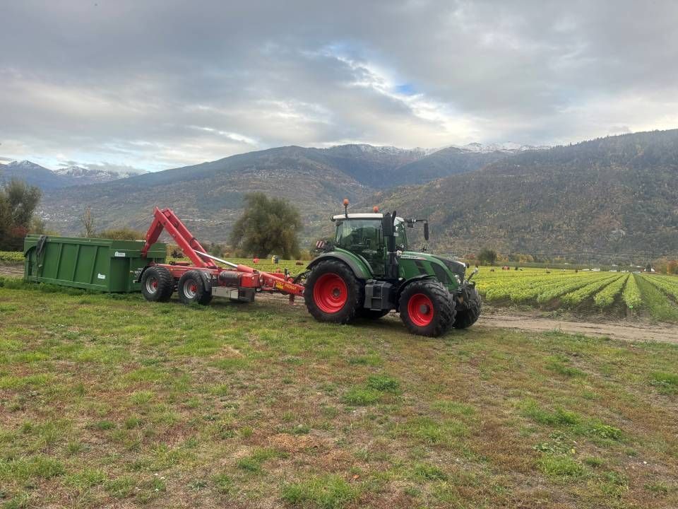 Un tracteur vert tire une benne à ordures verte dans un champ, avec des montagnes en arrière-plan.