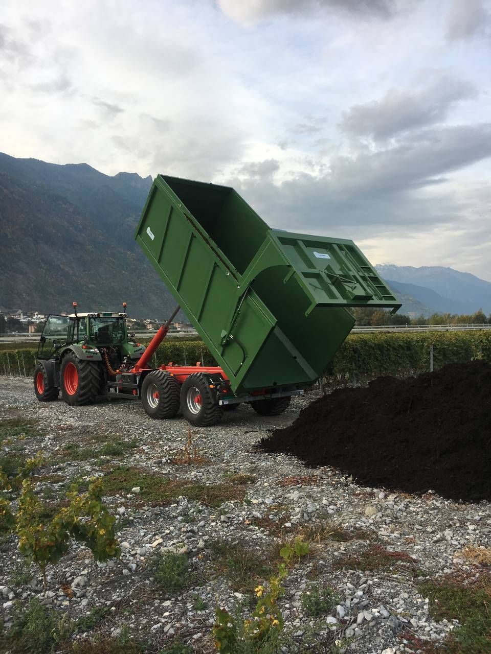 Un tracteur vert déverse de la terre noire en un tas. Montagnes et vignoble en arrière-plan.