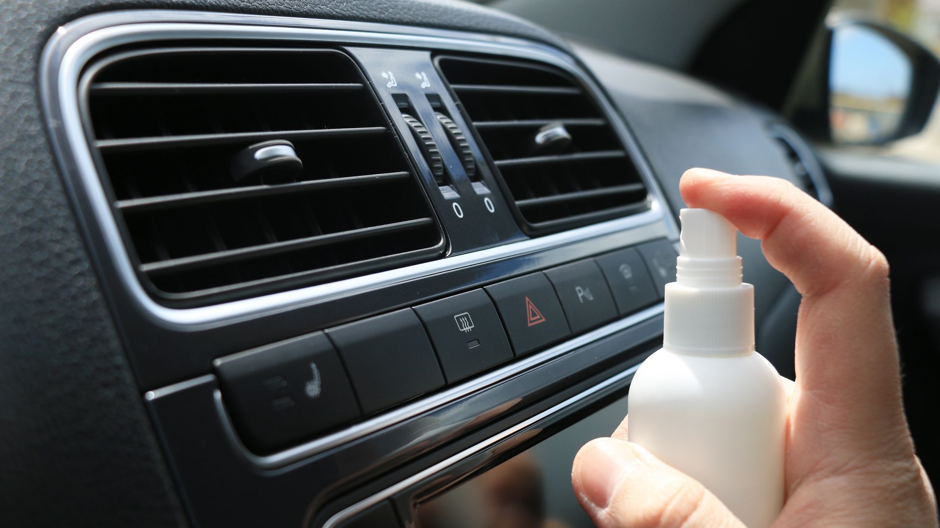 Main pulvérisant un flacon blanc vers les aérations du tableau de bord d'une voiture. Intérieur noir avec garnitures argentées.