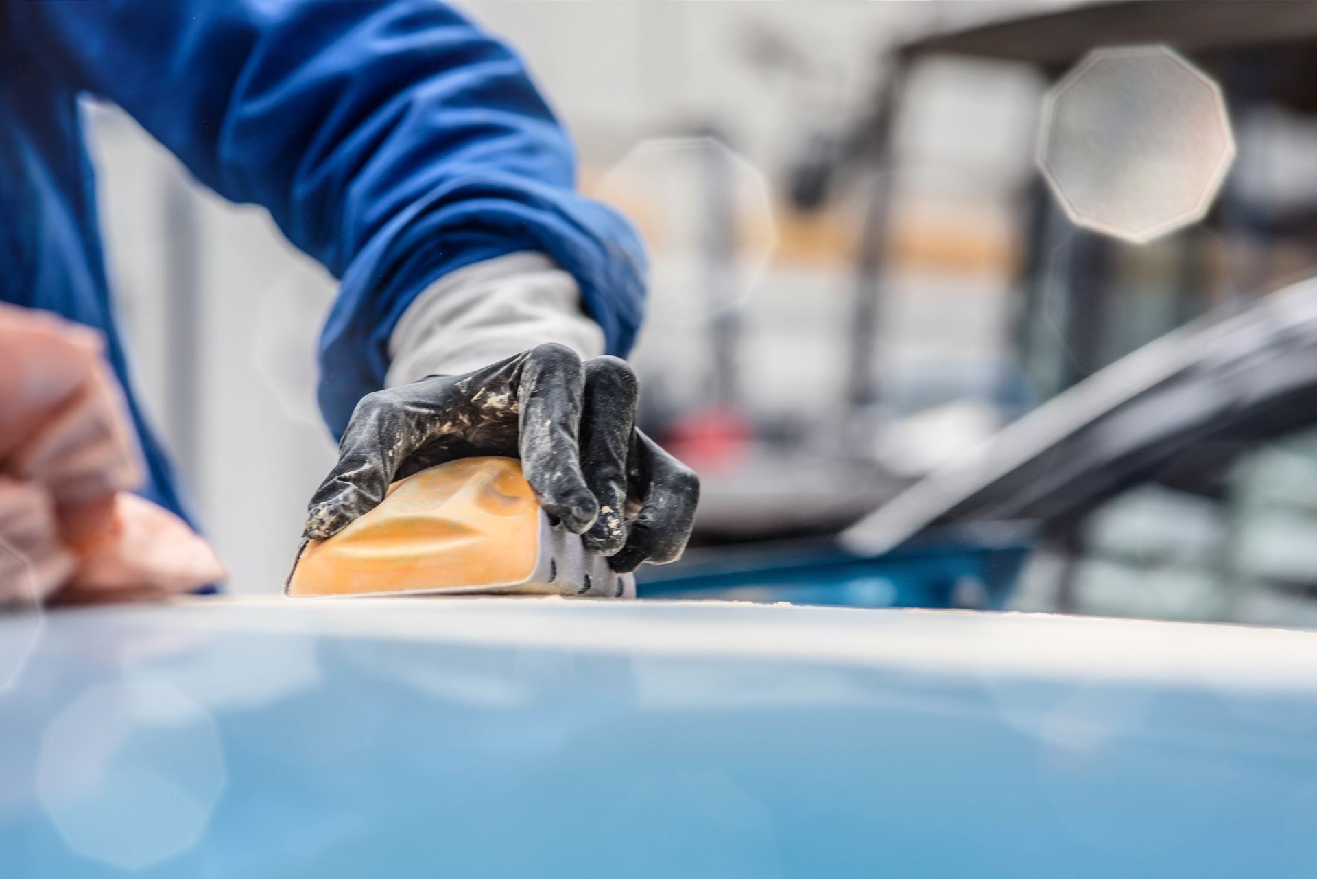 Une personne en combinaison de travail bleue ponce le capot d'une voiture et porte des gants.