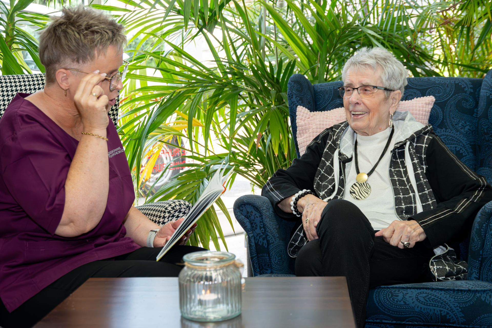 Two people seated on a patio in conversation, one holding a notebook, with plants and a candle on the table.