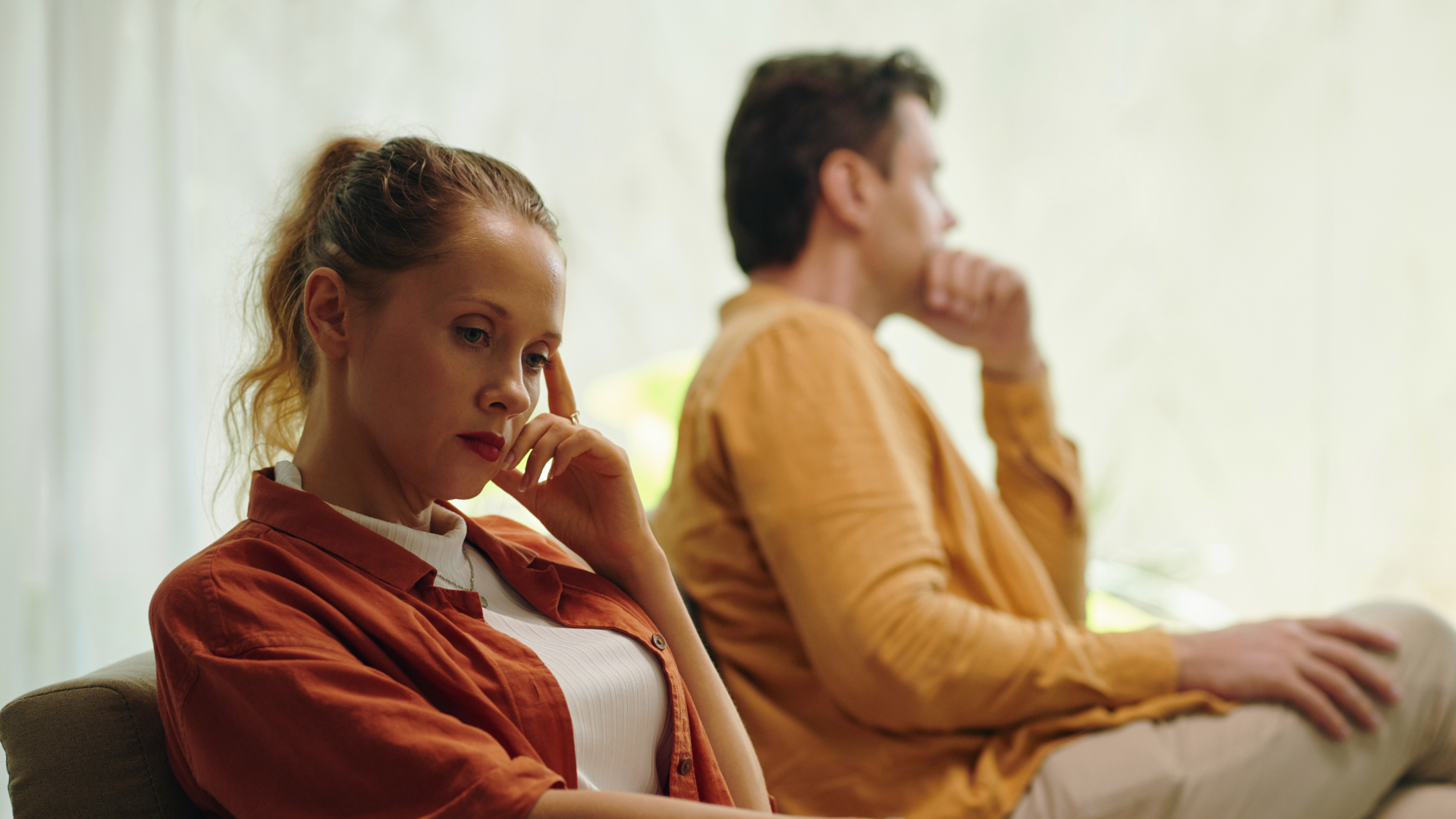 A person sitting in the foreground looks away with a serious expression, while another person sits turned away behind them.
