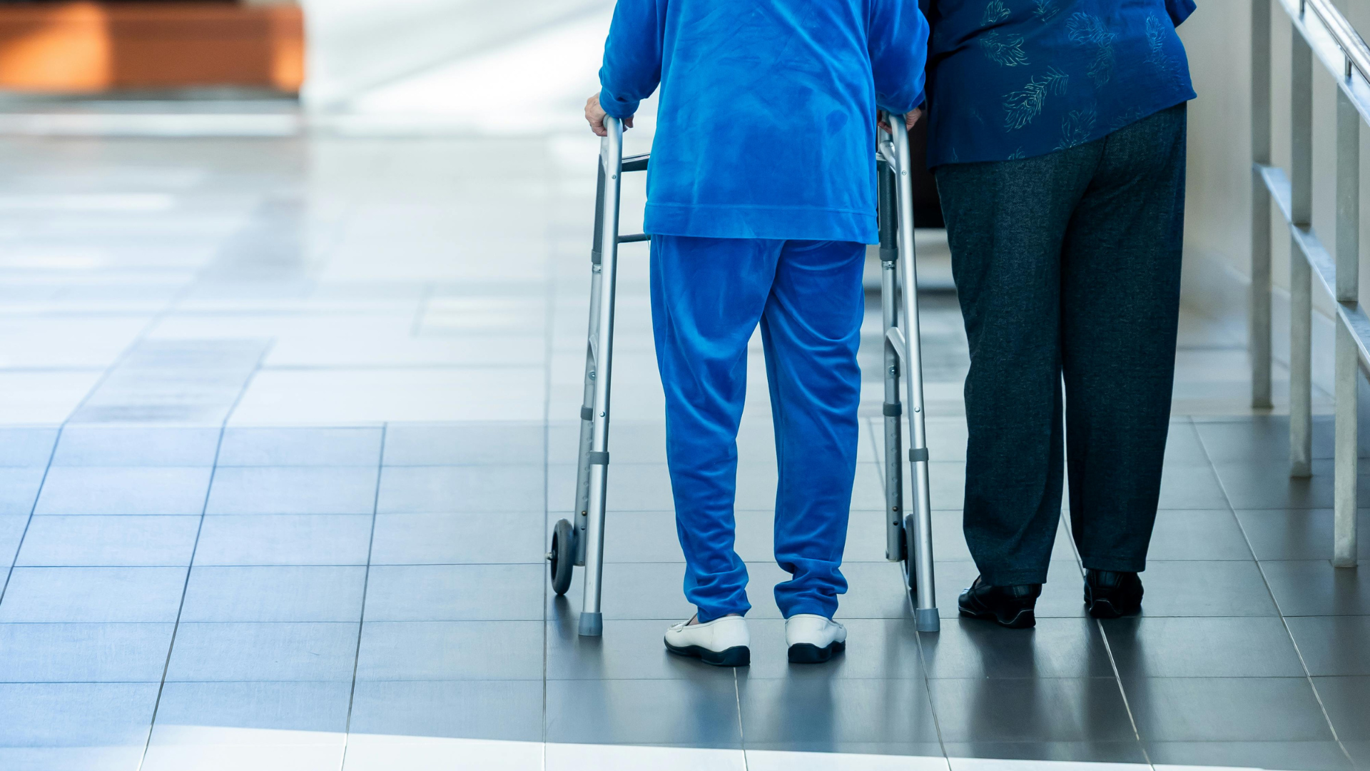 Zwei Personen gehen nebeneinander in einem hellen Flur, eine davon benutzt einen Rollator.