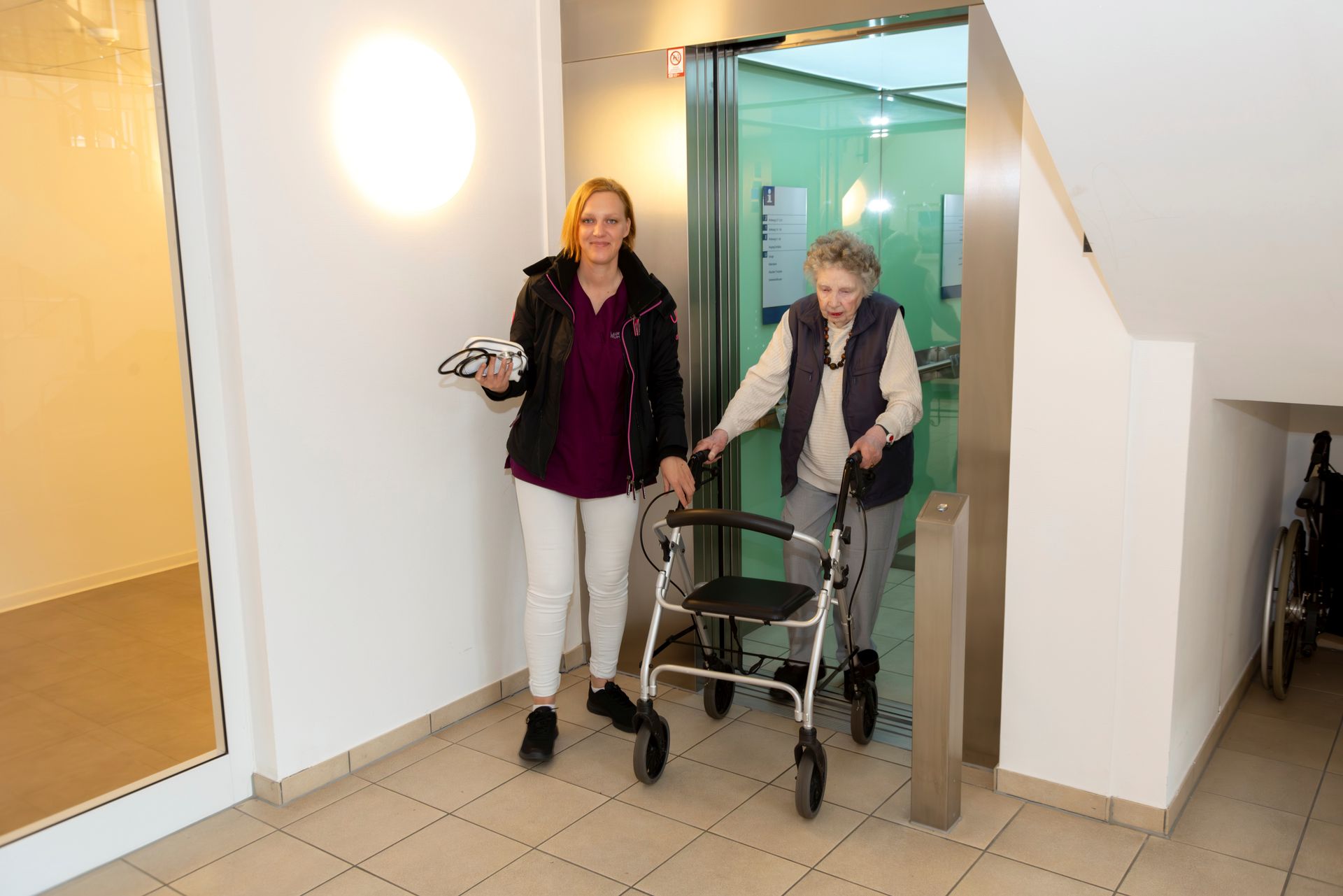 Alte Frau mit Rollator wird von Pflegerin us Fahrstuhl begleitet