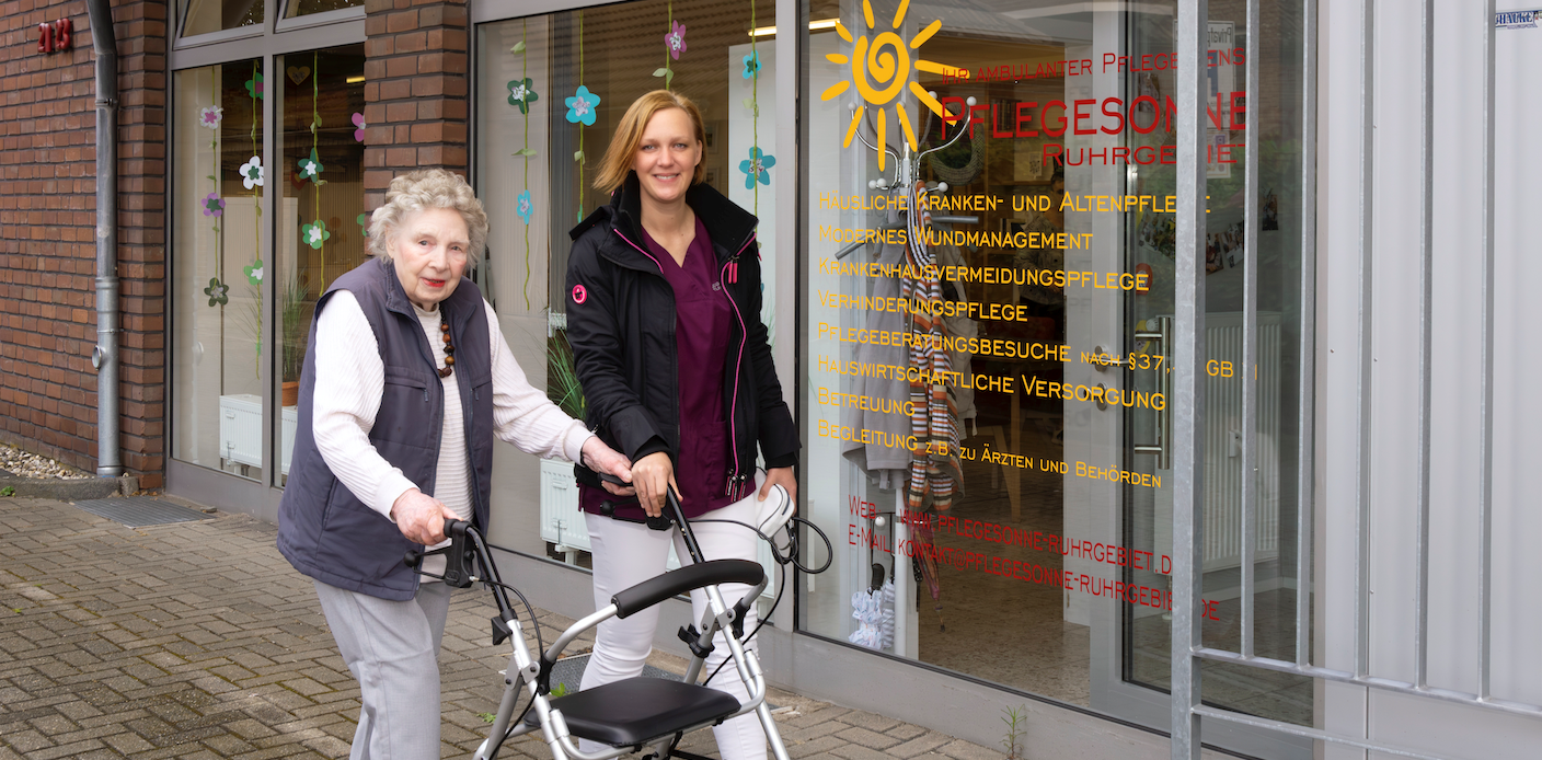 Seniorin mit Rollator und jüngerer Begleitung. vor dem Ladenlokal der Pflegesonne