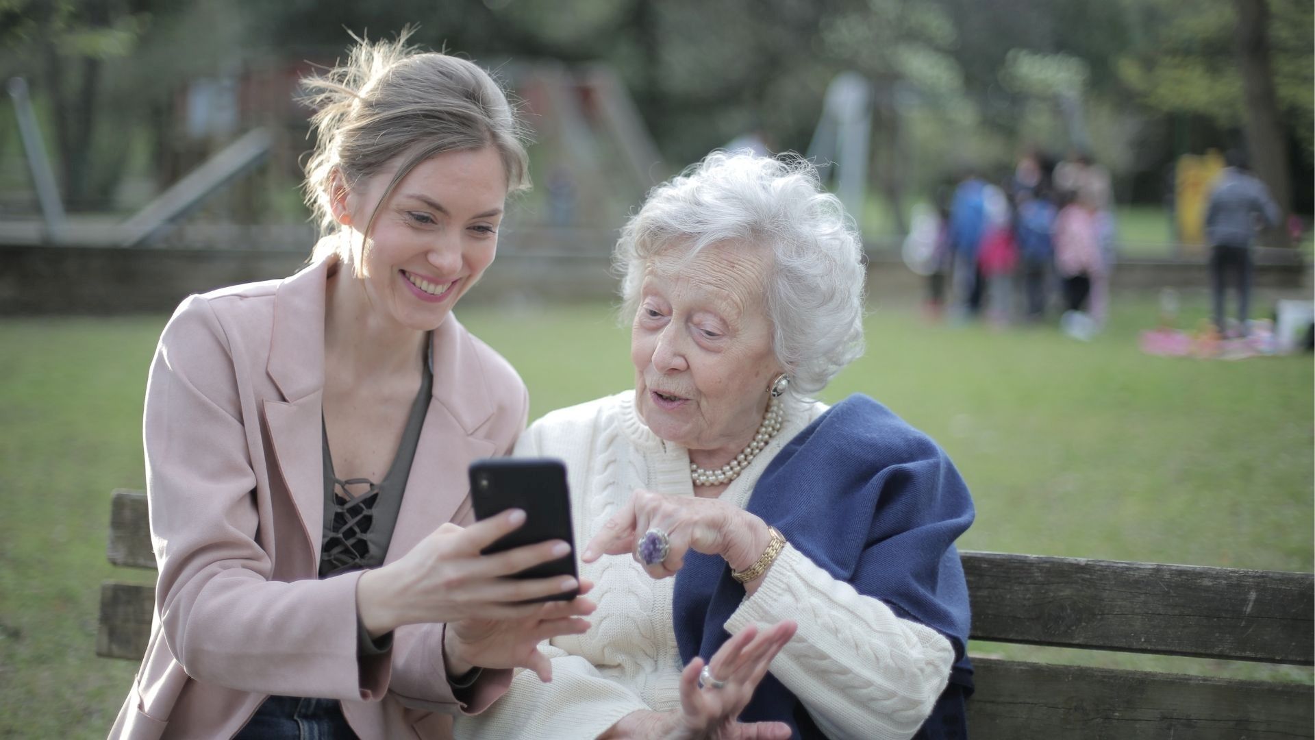 Junge Frau zeigt alter Frau etwas auf dem Smartphone, sie sitzen auf einer Bank im Park.