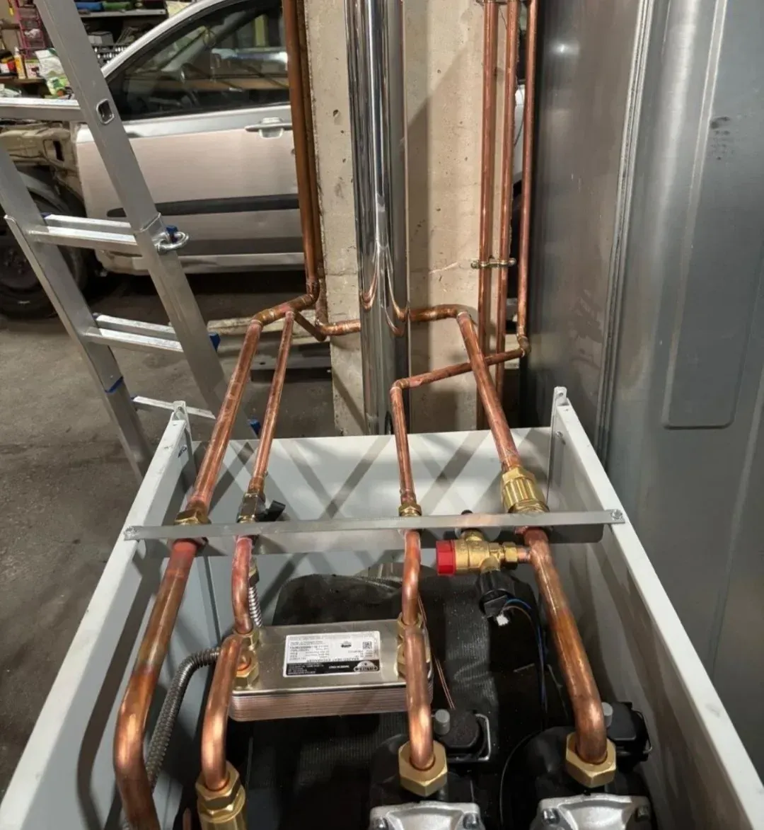 Tuberías de cobre y equipos de climatización dentro de un armario de servicios públicos con un coche al fondo.