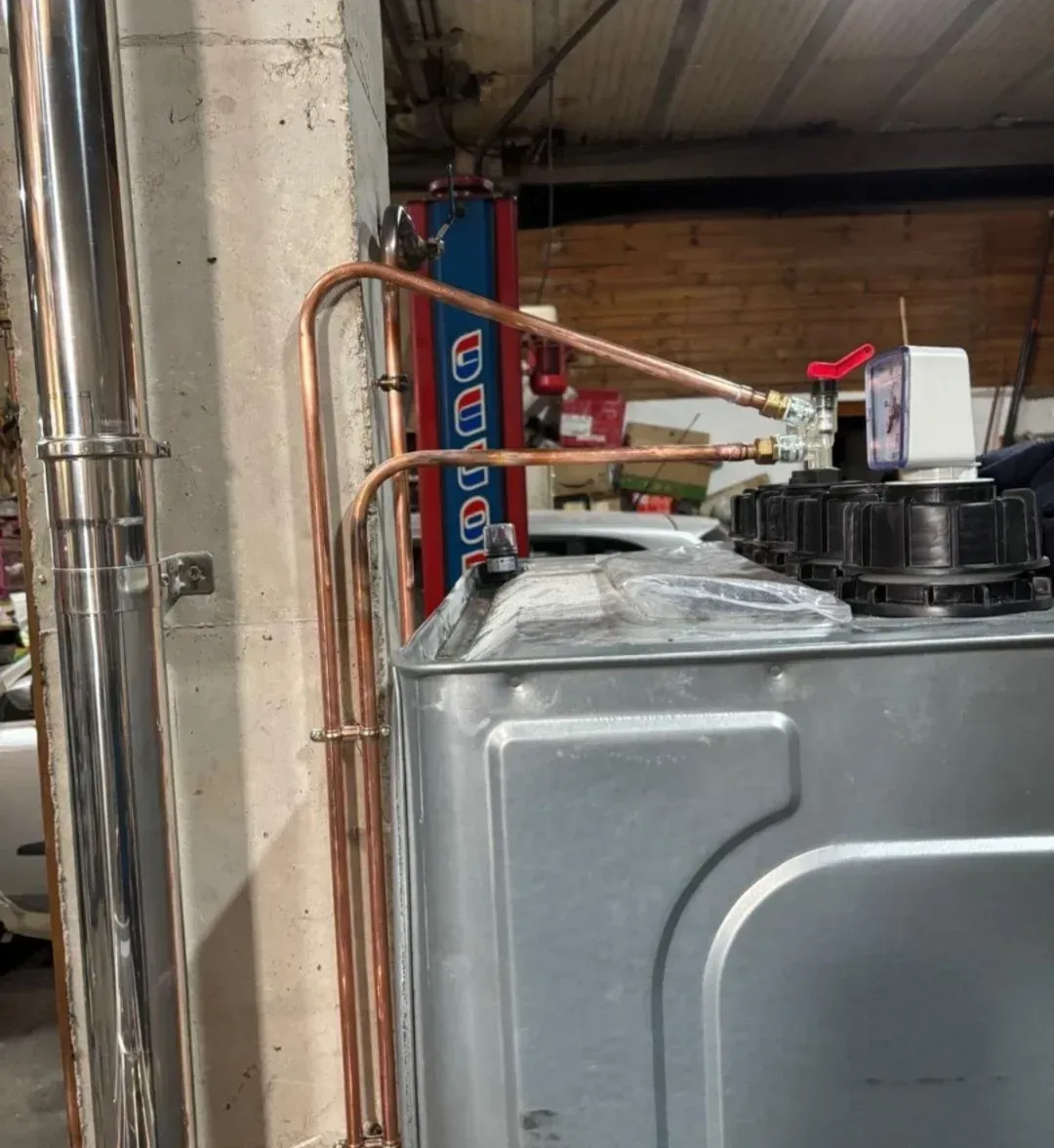 Tuberías de cobre conectadas a un tanque de metal en un garaje o cuarto de servicio, con una escalera roja al fondo.