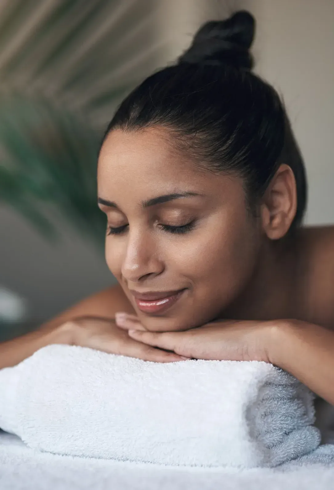 Mujer descansando su rostro sobre una toalla blanca doblada, con los ojos cerrados, sonriendo, en un entorno de spa.
