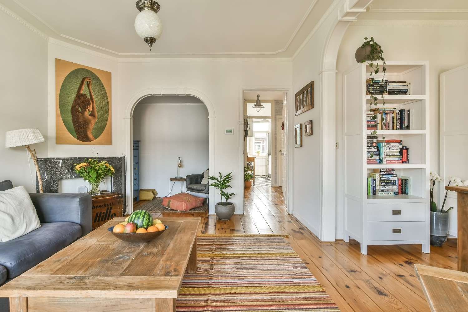 Sala de estar luminosa con sofá gris, mesa de centro de madera, alfombra a rayas, puerta arqueada y estantería blanca.