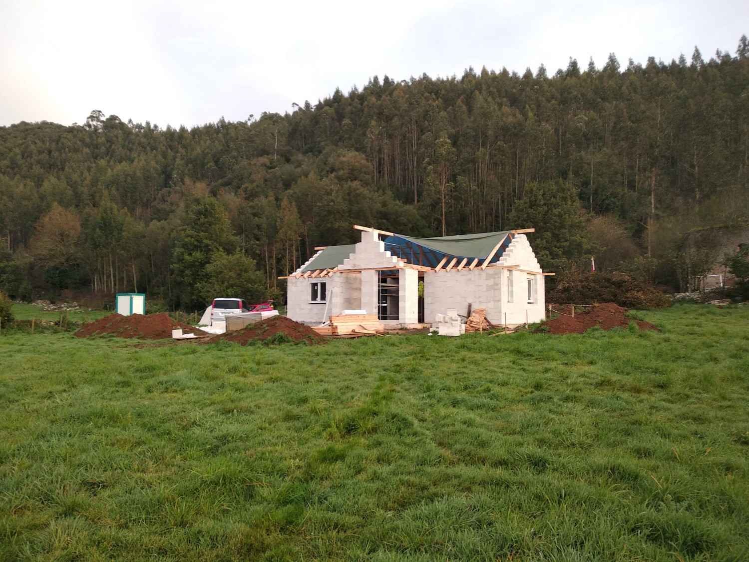 Casa blanca parcialmente construida en un campo verde con colinas boscosas detrás.