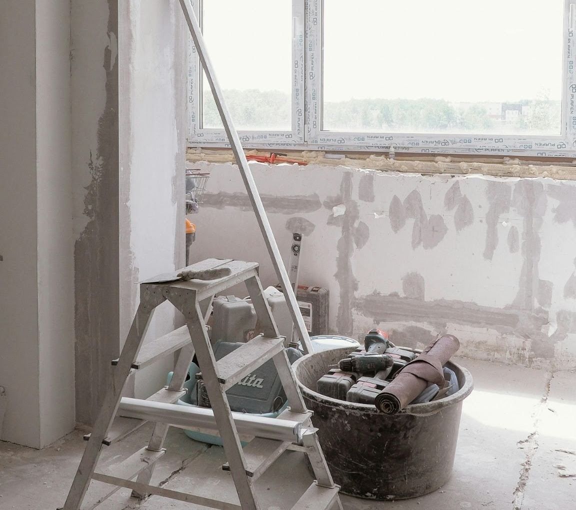 Cuarto de renovación con escalera de mano, cubo de pintura y paredes enlucidas irregulares cerca de una ventana.