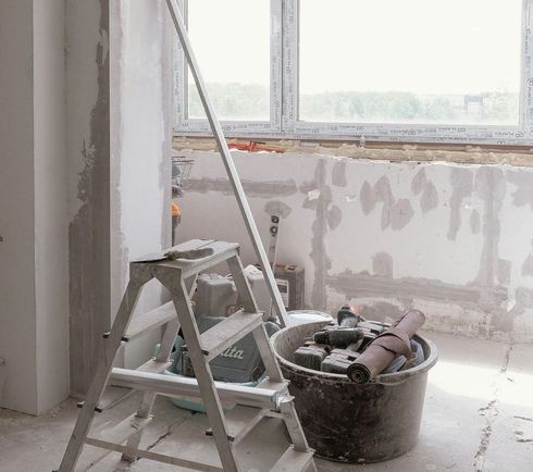 Cuarto de renovación con escalera de mano, cubo de pintura y paredes enlucidas irregulares cerca de una ventana.