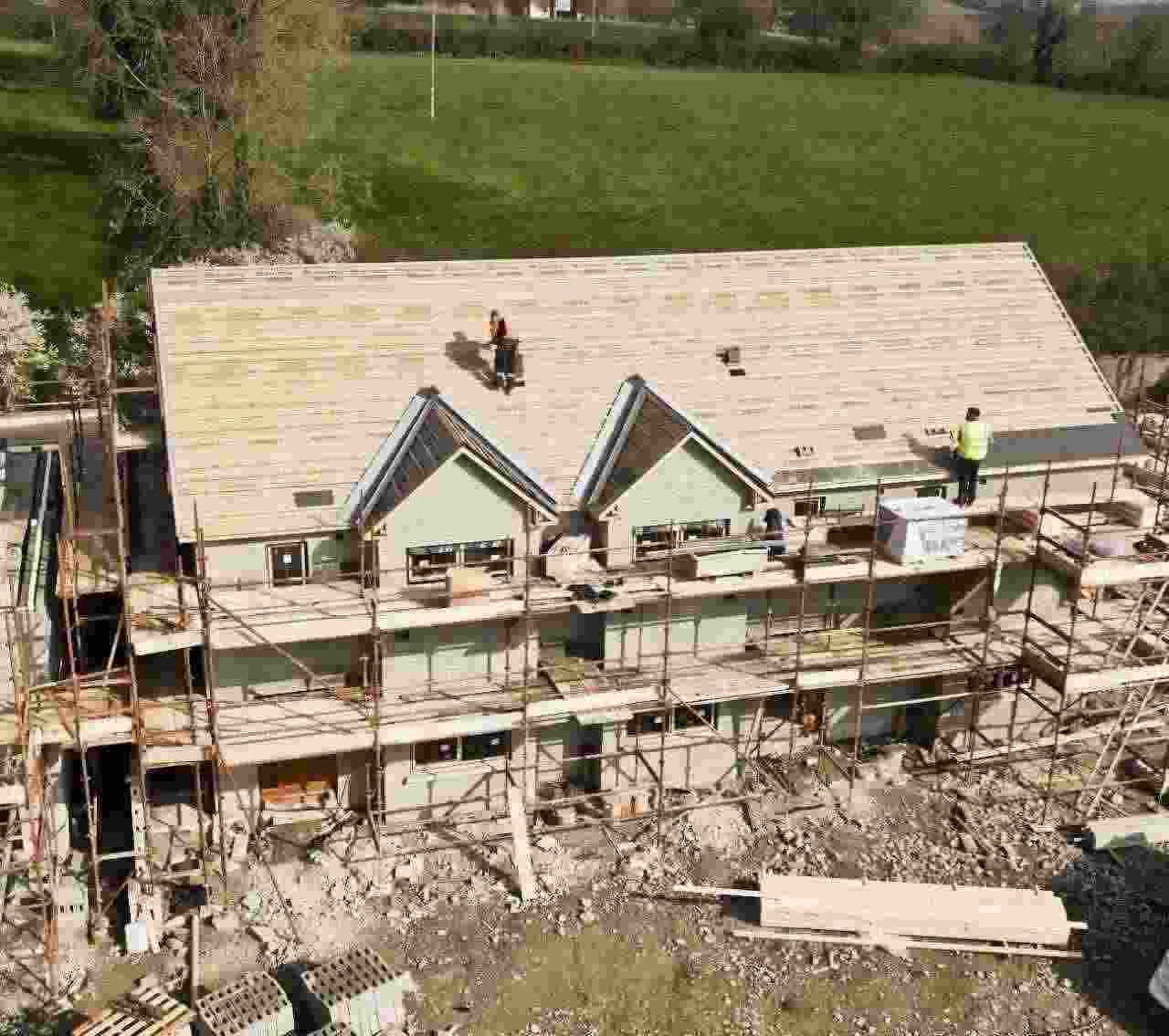 Trabajadores en el tejado de una casa en construcción, con andamios y un campo verde al fondo.