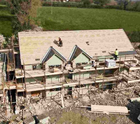 Trabajadores en el tejado de una casa en construcción, con andamios y un campo verde al fondo.