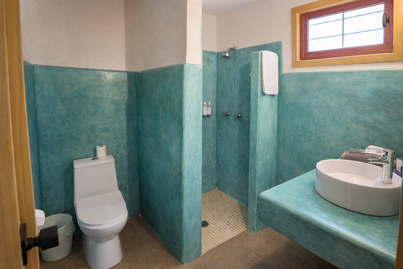 Un ba&ntilde;o con paredes enlucidas de color turquesa, un inodoro blanco, una ducha a ras de suelo y un lavabo blanco sobre una repisa empotrada.