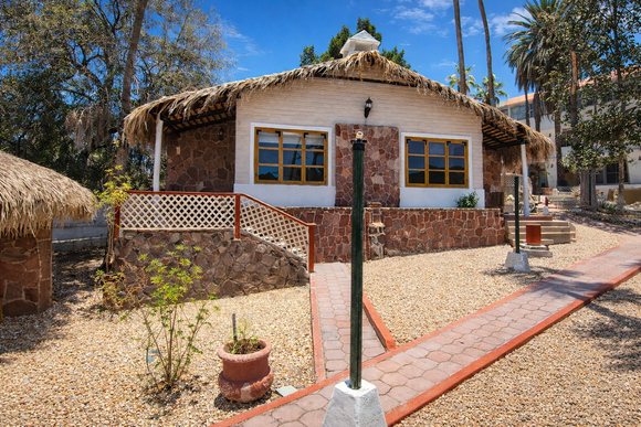 Una caba&ntilde;a de piedra con techo de paja y paredes de color tostado, situado en un paisaje soleado y des&eacute;rtico con un camino pavimentado.
