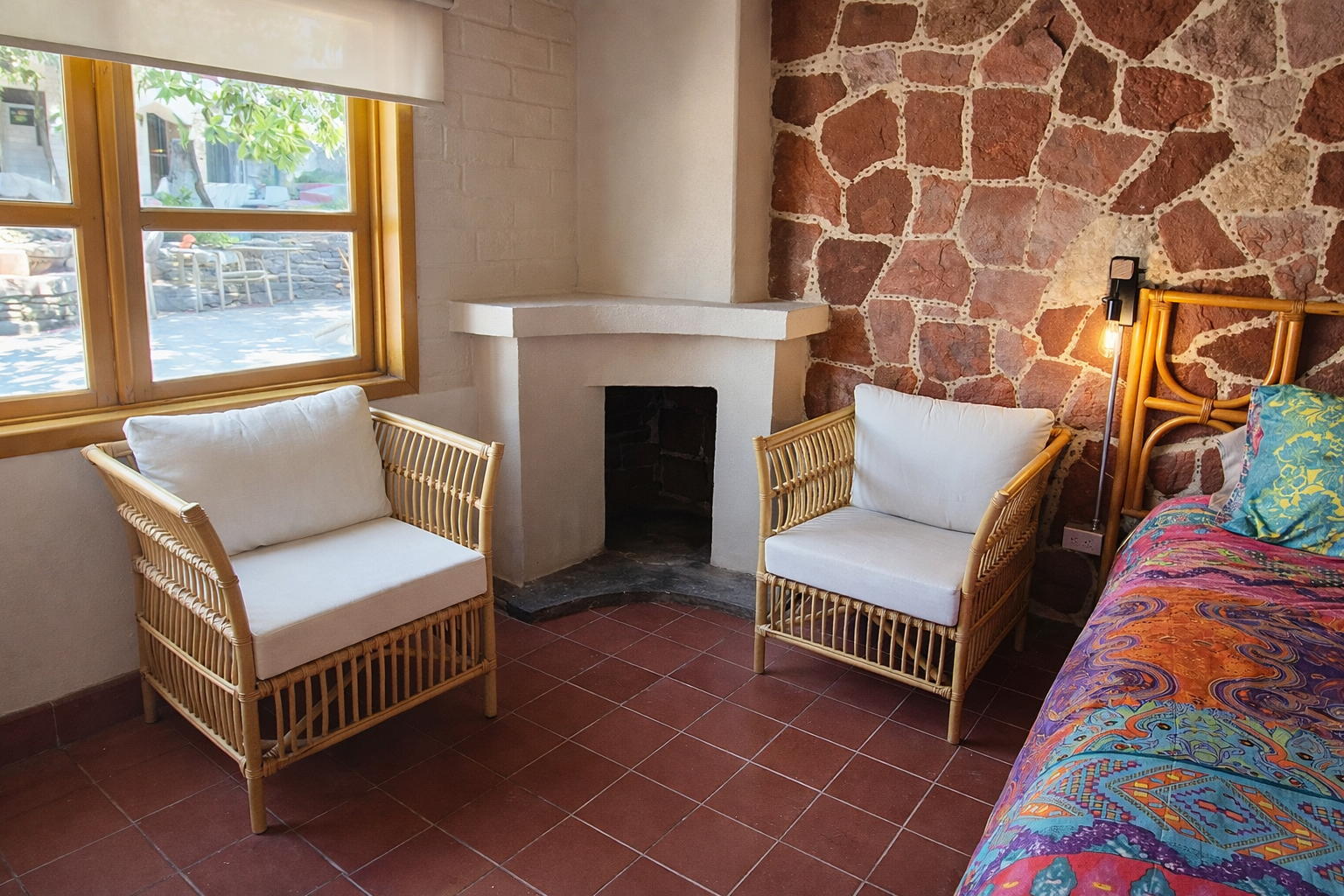 Una habitaci&oacute;n con dos sillas de mimbre, una chimenea en la esquina, una pared de piedra y una cama con una colcha colorida estampada.