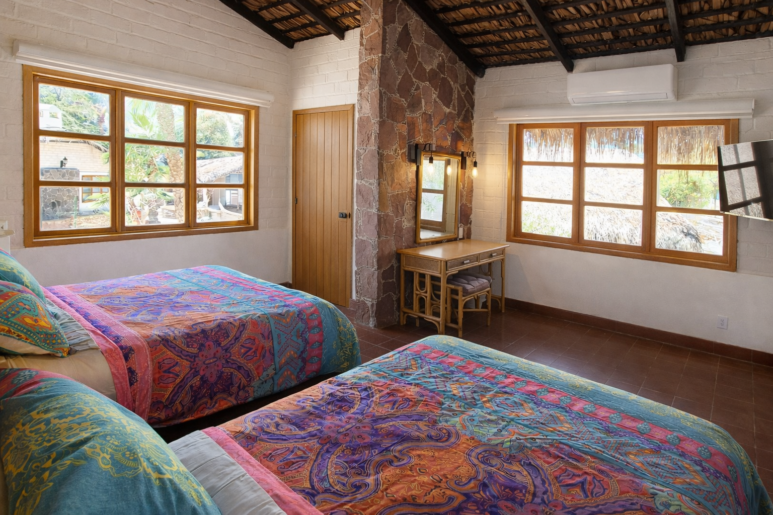Dos camas con coloridas colchas con estampado de cachemir en una habitaci&oacute;n con paredes blancas, ventanas con marcos de madera y una pared decorativa de piedra.