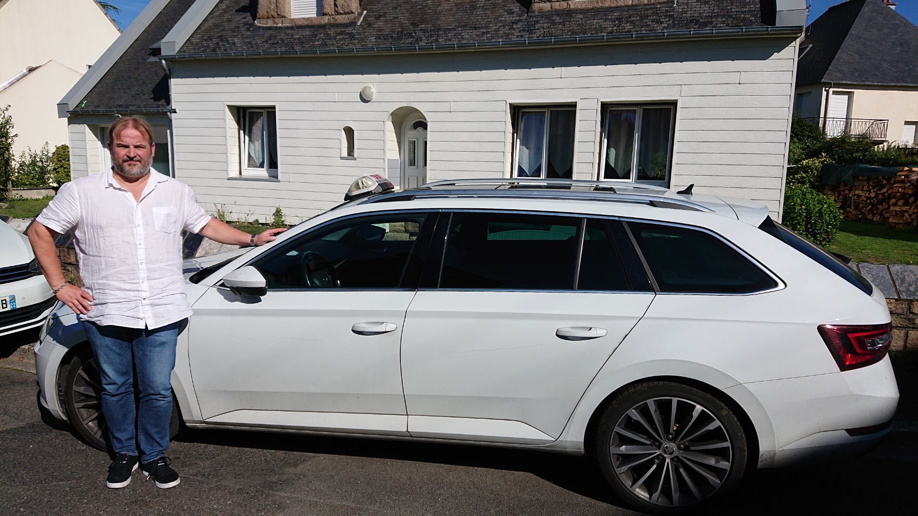 Un homme debout à côté d’un break blanc garé devant une maison, dans une rue ensoleillée.