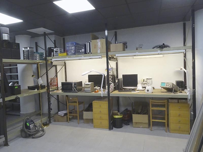 Taller con bancos de trabajo, estanterías para equipos y dos computadoras. Luces fluorescentes iluminan el espacio de trabajo.