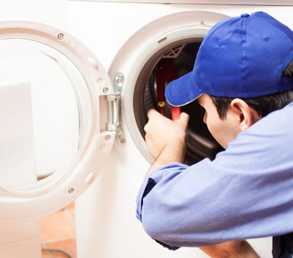 Un hombre con gorra azul repara una lavadora, utilizando una herramienta dentro del tambor.