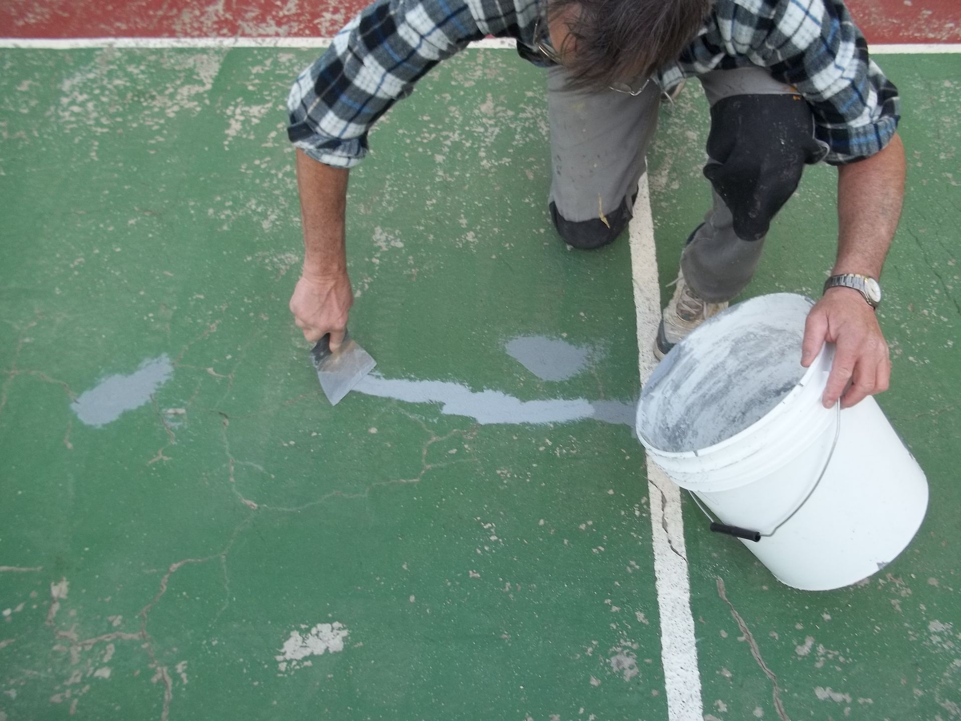 Une personne applique un enduit de rebouchage gris sur un terrain vert, tenant un seau et une truelle.