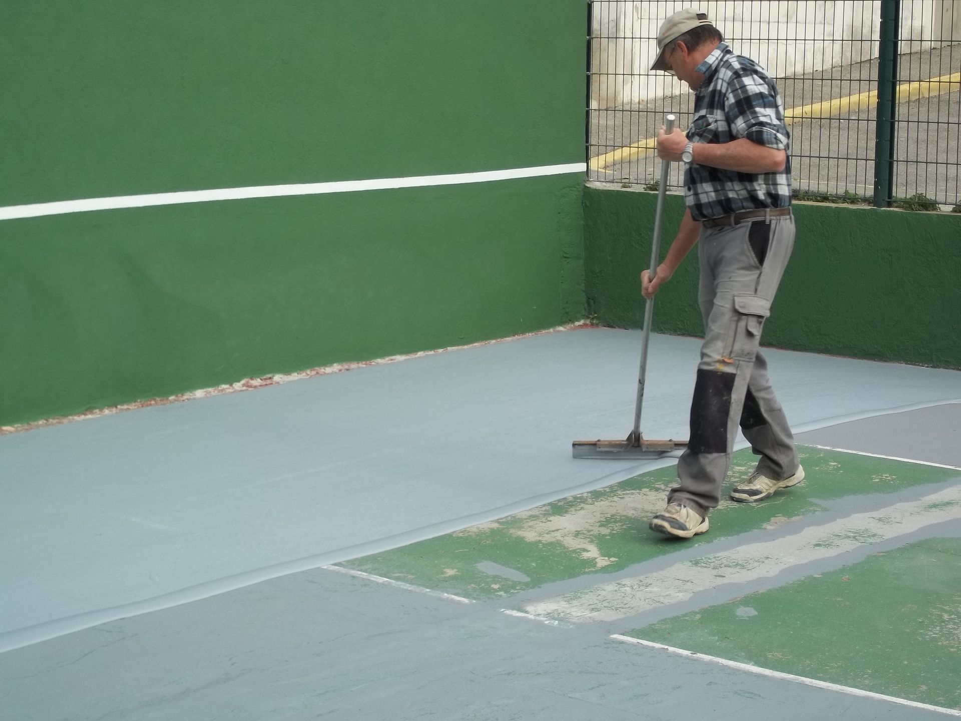 Un homme portant une casquette, une chemise et un pantalon de travail utilise une raclette sur la surface peinte d'un terrain de sport.
